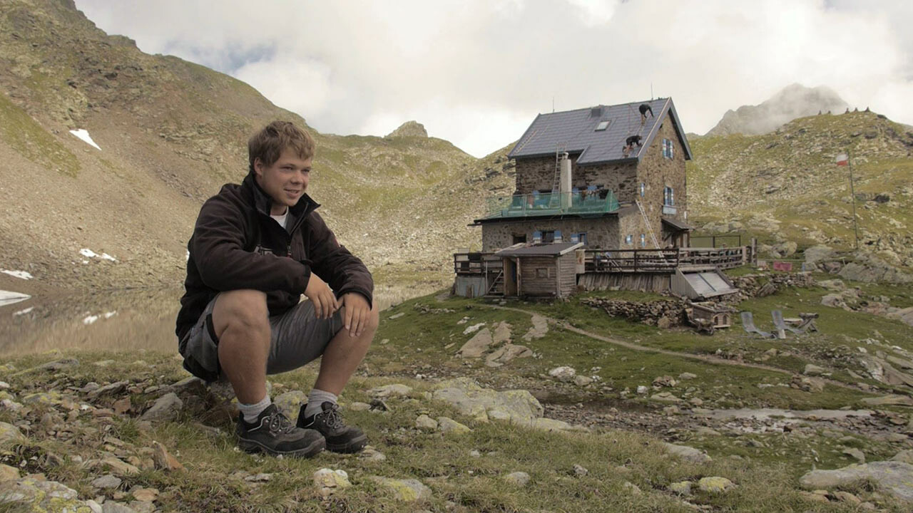 Lärlingen Jakob Schöttl sitter på en sten. I bakgrunden syns ”flaggskeppet Schartenhütte” som klätts med PREFA takplatta. 