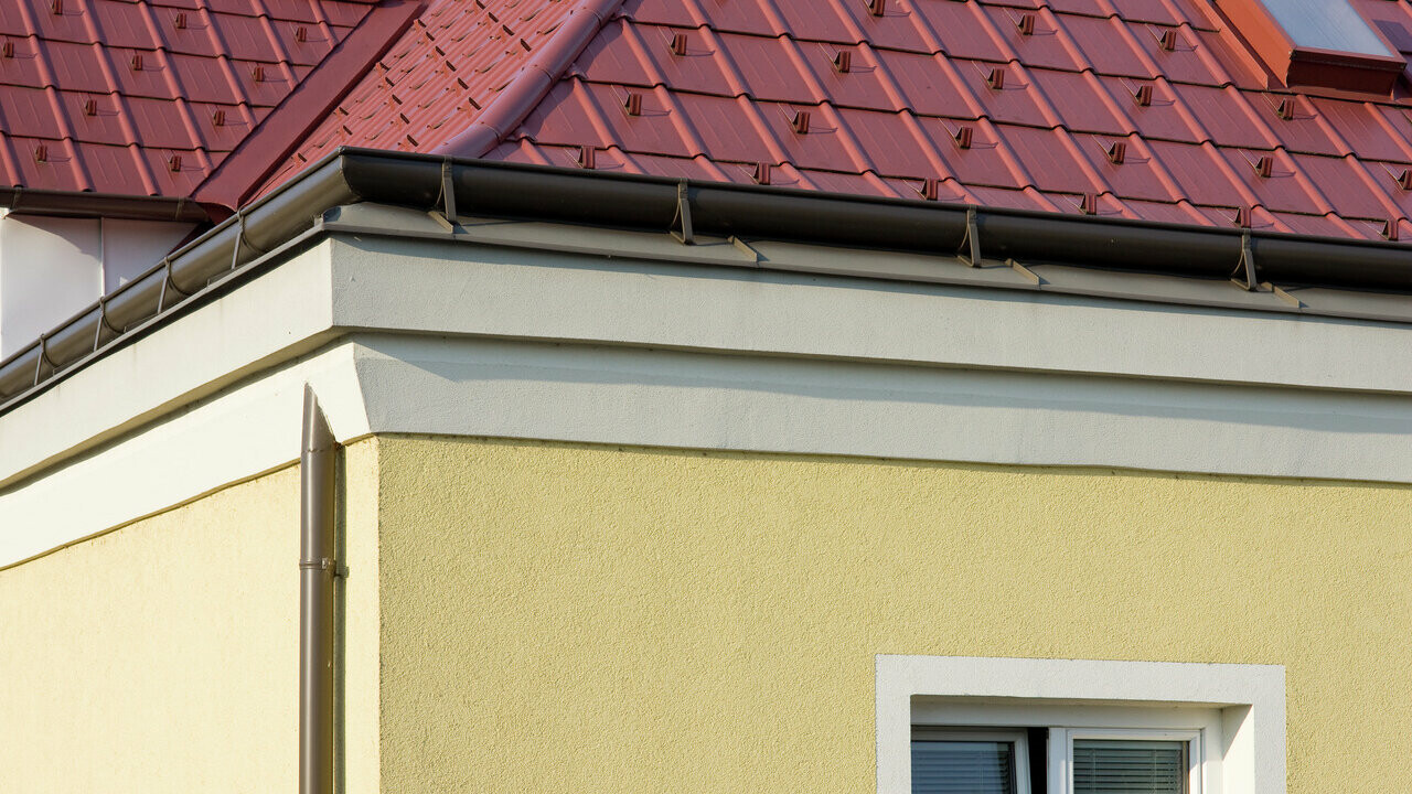 Hörn på hus med gul putsfasad och PREFA fotränna i brunt med röda PREFA takplattor på taket
