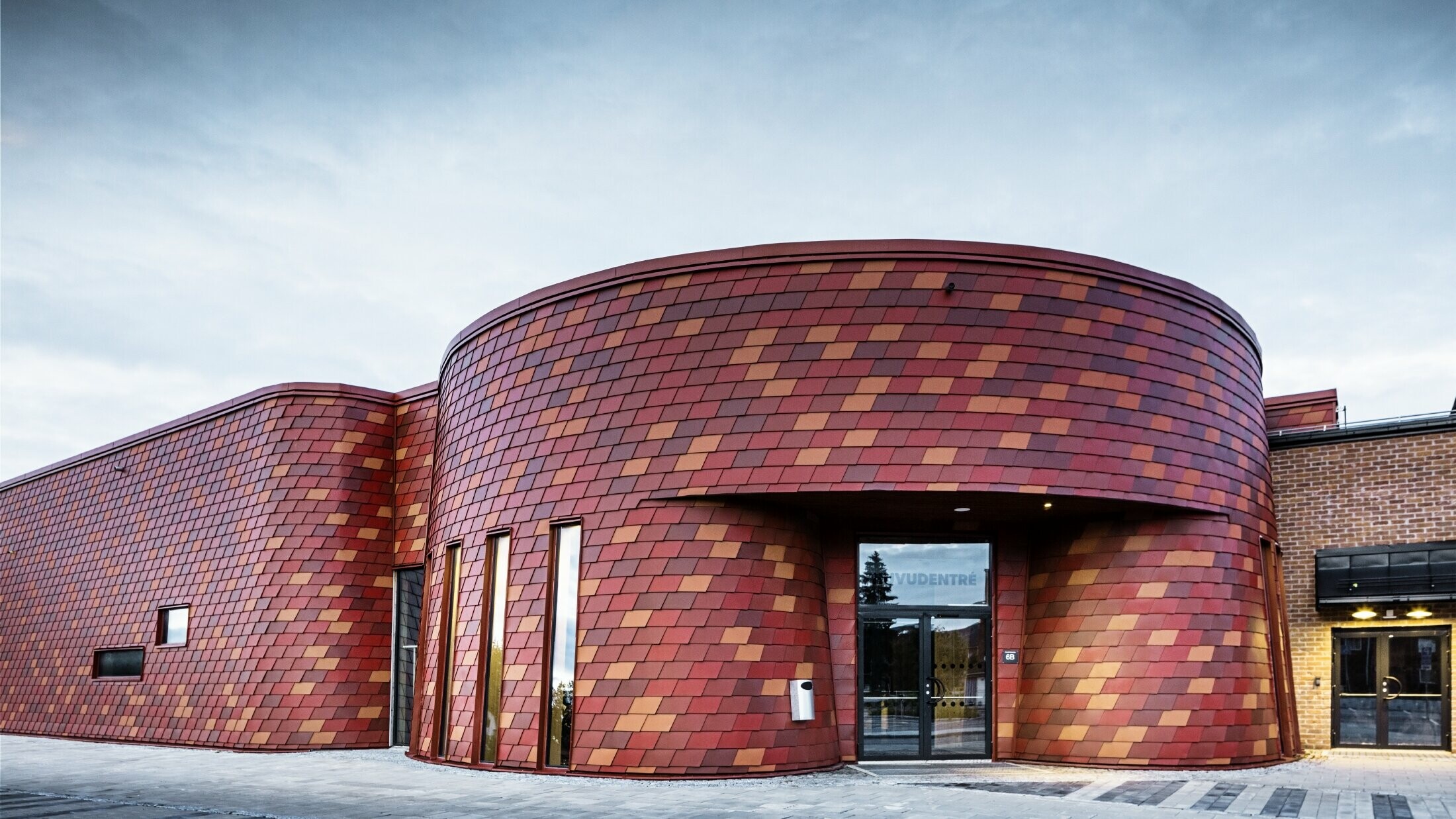 Ishall med flackt tak och svängd fasad – fasaden är täckt med väggshinglar i aluminium från PREFA i olika röda nyanser: oxidröd, tegelröd, rödbrun, brunröd