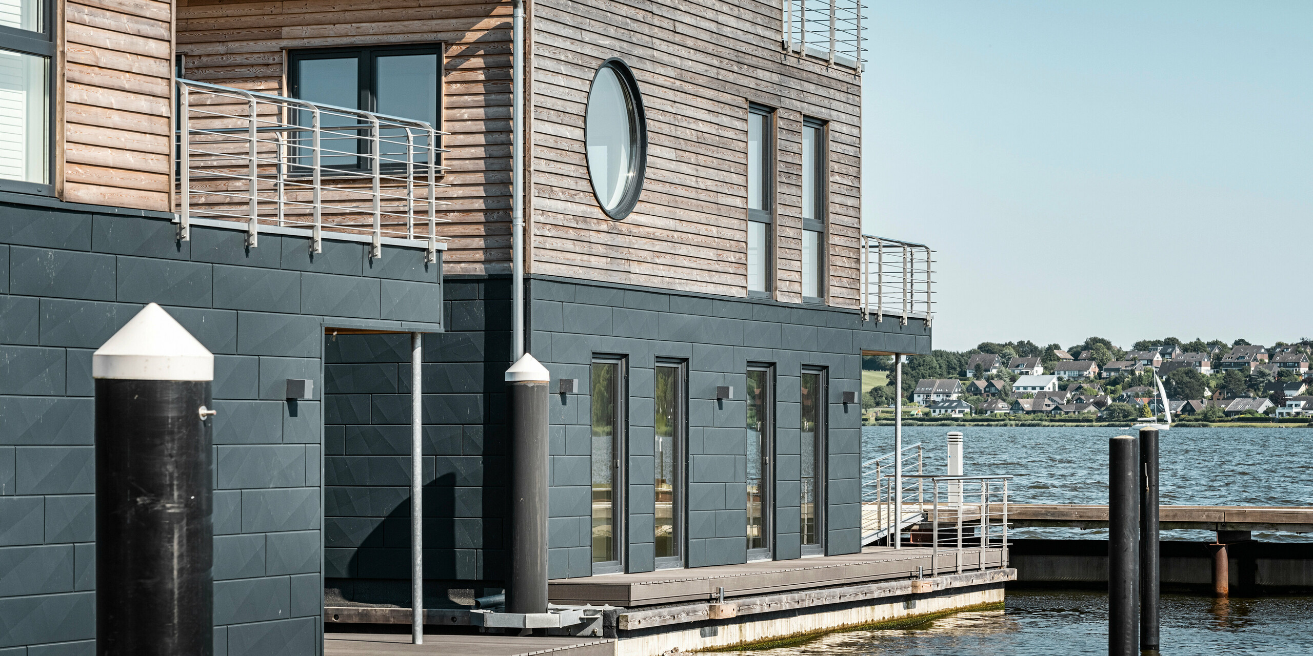 Nahaufnahme eines stilvollen Wasserhauses im Pionierhafen Schleswig mit einer Fassade aus PREFA Siding.X in P.10 Anthrazit und natürlichem Holz am oberen Stockwerk. Ein rundes und mehrere eckige Fenster sowie klare Linien an der Konstruktion bieten einen modernen Kontrast zur traditionellen Landschaft im Hintergrund. Diese schwimmenden Häuser sind mit ihren nachhaltigen Materialien und direktem Zugang zum Wasser ausgestattet. Die elf Wasserhäuser in Schleswig sind ein Beispiel für modernes Wohnen in Harmonie mit einer maritimen Umgebung.