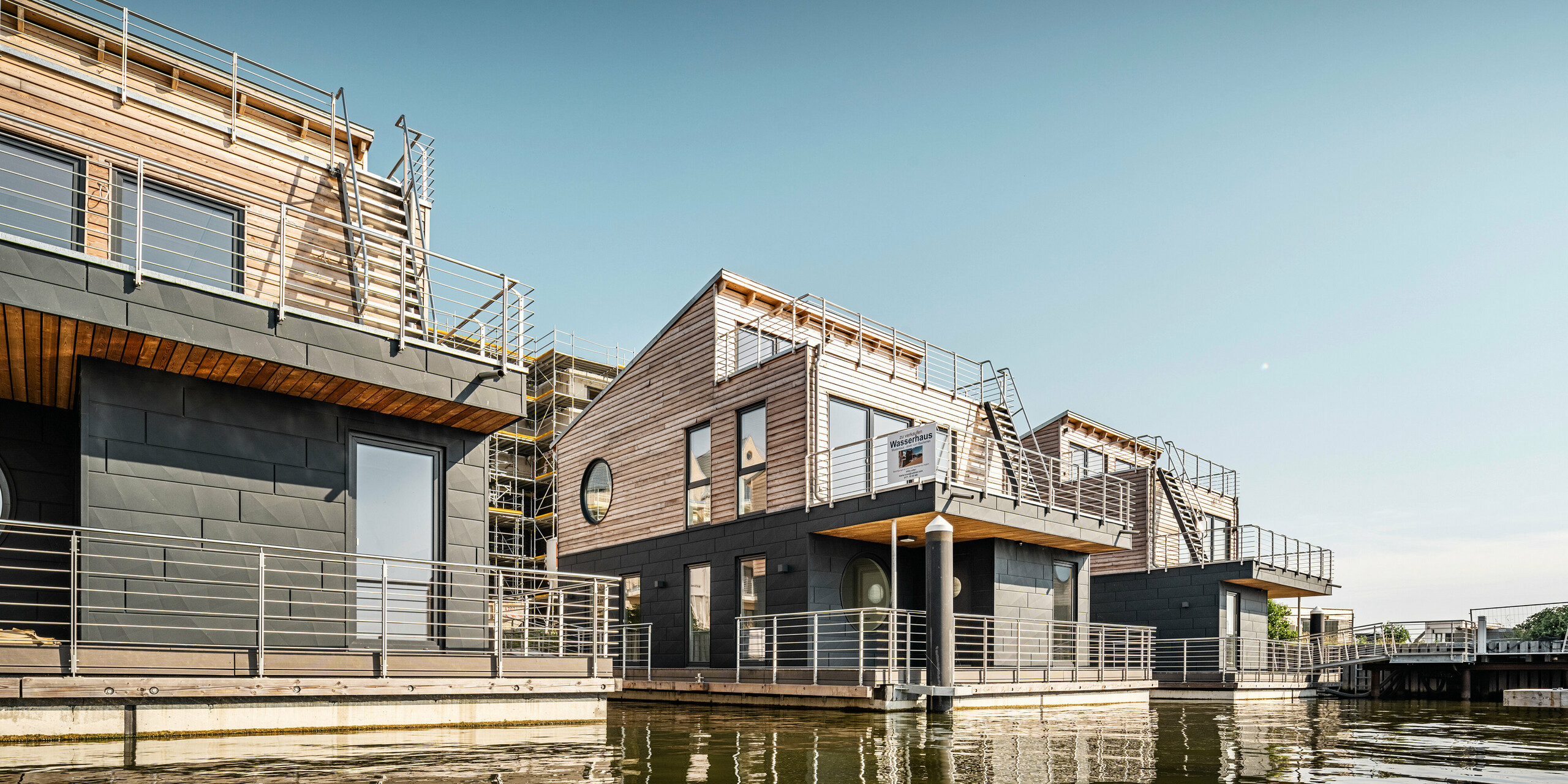 Eindrucksvolle Ansicht moderner Wasserhäuser im Pionierhafen Schleswig, die mit einer Kombination aus PREFA Siding.X in P.10 Anthrazit und horizontalen Holzpaneelen ausgestattet sind, was eine ansprechende Verbindung zwischen moderner Architektur und natürlichen Materialien schafft. Die Gebäude stehen auf Schwimmkörpern, die eine dauerhafte und stabile Lage im Wasser garantieren, und sind mit großzügigen Dachterrassen versehen, die einen umfassenden Blick auf die umgebende Wasserlandschaft ermöglichen.