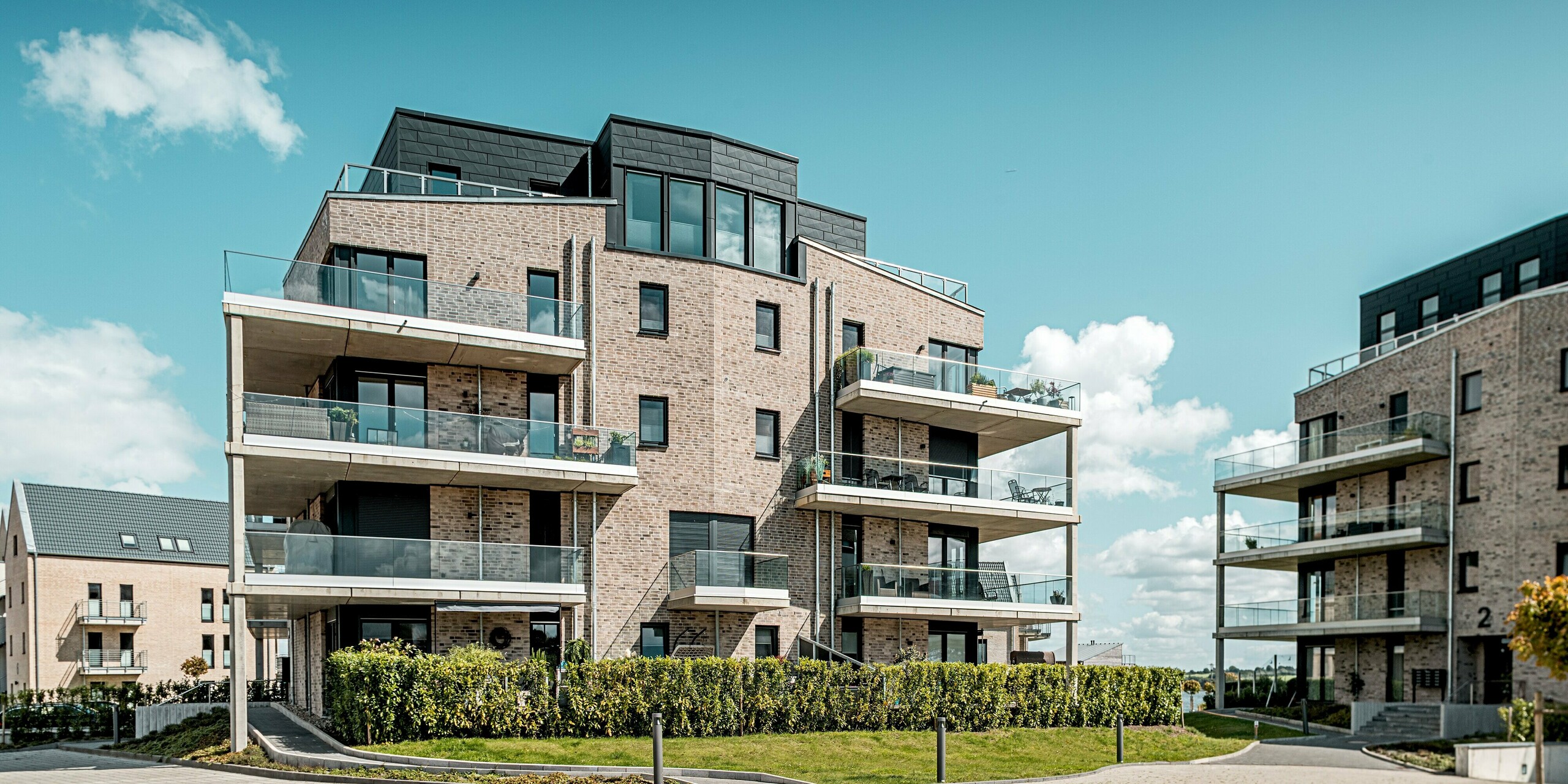 Die Wohnhausanlage „Hafen zur Freiheit“ präsentiert sich in einer offenen Hofsituation, bei der sich moderne Klinkerarchitektur harmonisch mit den markanten PREFA Siding.X Akzenten in P.10 Anthrazit verbindet. Das zentrale Gebäude mit seinen auskragenden Balkonen und klaren Linien steht im Mittelpunkt und wirkt durch die dynamisch strukturierten Aluminiumflächen besonders kraftvoll. Großzügige Glasbrüstungen verstärken den modernen Look und reflektieren das Tageslicht. Die langlebigen, korrosionsbeständigen PREFA Fassadenpaneele bieten optimalen Schutz vor den rauen Küstenbedingungen und unterstreichen die hochwertige Bauqualität.