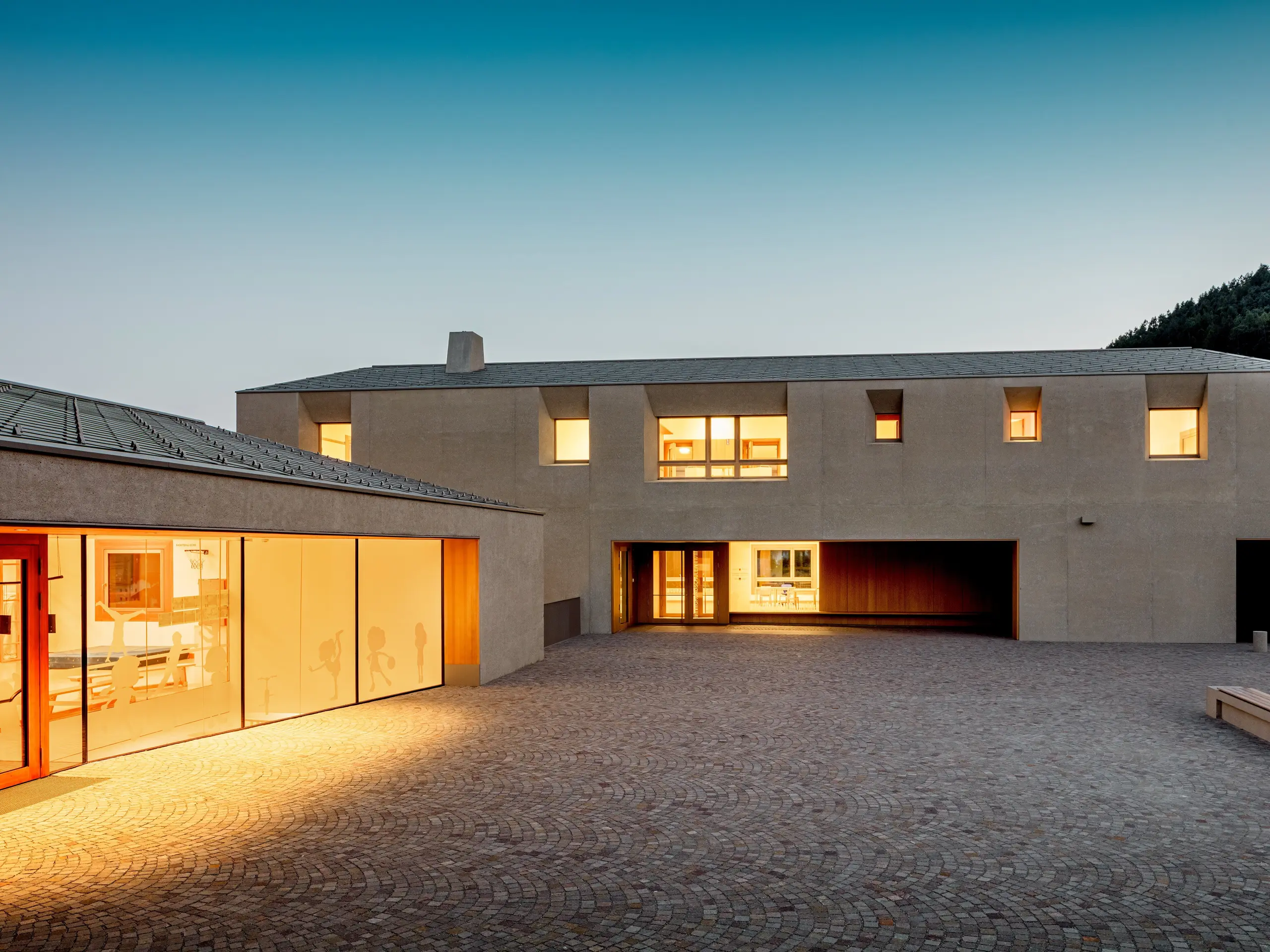 In der Abenddämmerung leuchtet das Bildungszentrum Riffian warm über den gepflasterten Schulhof. PREFA Dachrauten 44 × 44 in P.10 Bronze krönen Kindergarten und Volksschule mit präziser Aluminiumdeckung, verbinden klare Architektur mit wetterfester Langlebigkeit und setzen einen eleganten, ruhigen Akzent vor dem Südtiroler Berghang.