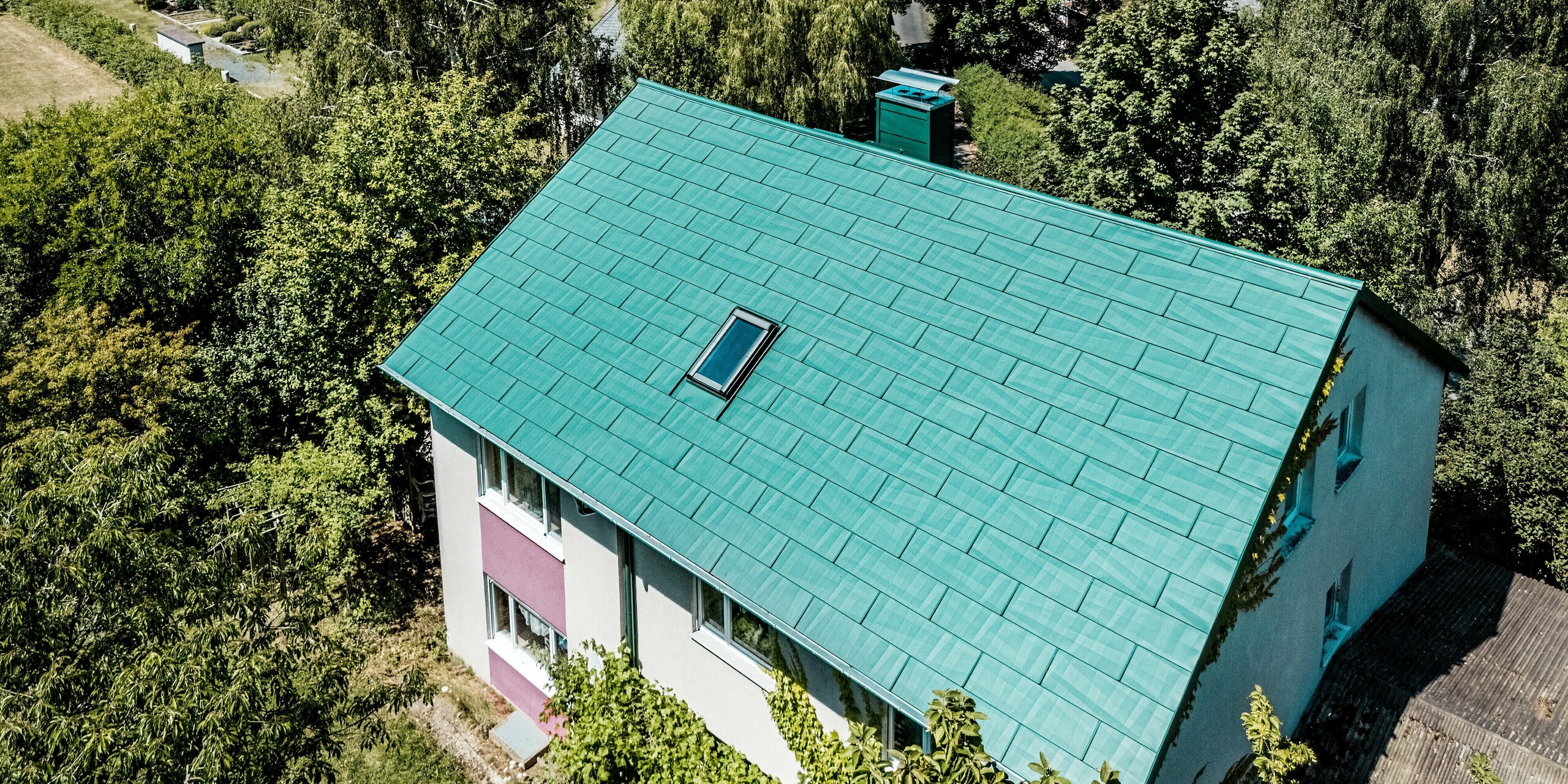 Modernisiertes Einfamilienhaus in Schwarzenbach-Martinlamitz mit moosgrünem PREFA Dachpaneel FX.12. Die Luftaufnahme zeigt das präzise verlegte Aluminiumdach mit charaktervoller Struktur und integriertem Dachfenster, eingebettet in eine grüne Umgebung. Die auffällige Farbkombination aus moosgrünem Dach und violett-weißer Fassade verleiht dem Haus eine unverwechselbare Optik. Das PREFA Dach überzeugt mit Langlebigkeit, Witterungsbeständigkeit und wartungsfreiem Komfort – ideal für stilvolle Sanierungen mit höchstem Qualitätsanspruch.