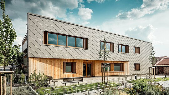 Die frontal aufgenommene Ansicht der Kindertagesstätte in Alzenau zeigt den einladenden Haupteingang mit seiner harmonischen Kombination aus bronzefarbenen PREFA Wandrauten 44 × 44 und vertikaler Holzfassade im Erdgeschoss. Die klar strukturierte Aluminiumverkleidung erzeugt ein elegantes Schuppenmuster, das durch großzügige Fenster mit Holzrahmen und vertikalen Lamellen rhythmisch unterbrochen wird. Im Vordergrund unterstreichen barrierefreie Wegeführung, Blumenbeete und Sitzbänke die hohe Aufenthaltsqualität. Das Zusammenspiel aus langlebigem Aluminium, natürlichem Holz und durchdachter Freiraumgestaltung macht das Gebäude zu einem architektonischen Vorzeigeprojekt für nachhaltige, funktionale und ästhetisch hochwertige Bildungsbauten mit PREFA Fassadensystemen.