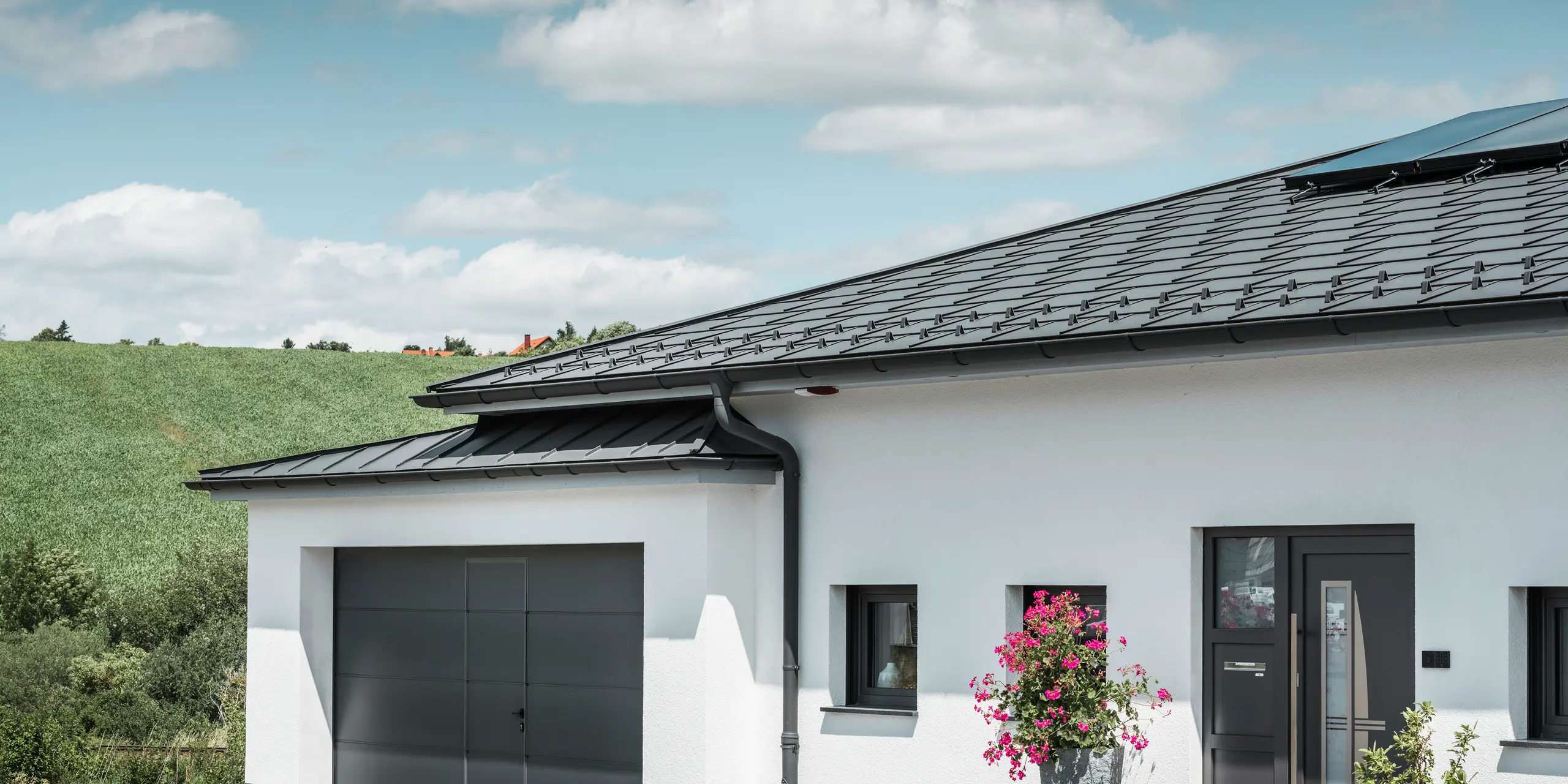 Bilden visar framsidan av en villa i Ilmenau, Thüringen, med ett elegant PREFA-tak i P.10 mörkgrå. Takplatta R.16 ger huset ett modernt utseende, medan det integrerade PREFA takavvattningssystemet med sin mörkgrå hängränna och avloppsrör garanterar effektiv avrinning av regnvatten. Garaget, utrustat med mörkgrå PREFALZ, smälter harmoniskt in i helhetsintrycket och understryker kvaliteten och stilen hos PREFA aluminiumprodukter.