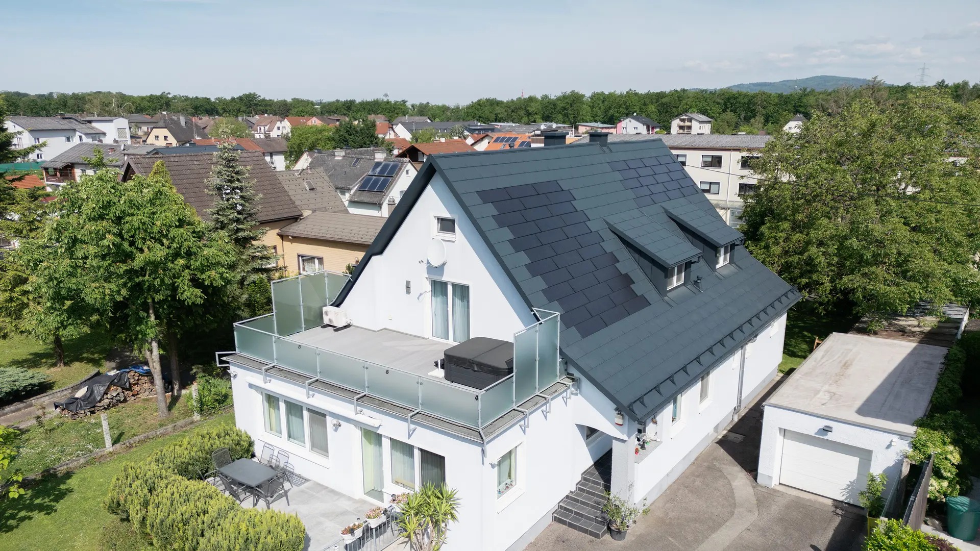 Fågelperspektiv på en villa efter takrenoveringen. Taket är nu täckt med stora PREFA-solar takplattor och PREFA-takplattor av typen R.16 i färgen P.10 antracit. Solar takplattorna är jämnt fördelade och skapar en modern, hållbar lösning. Byggnaden ger nu ett modernt och miljövänligt intryck. Balkongen i förgrunden har glasräcke och ligger på byggnadens översta våning.