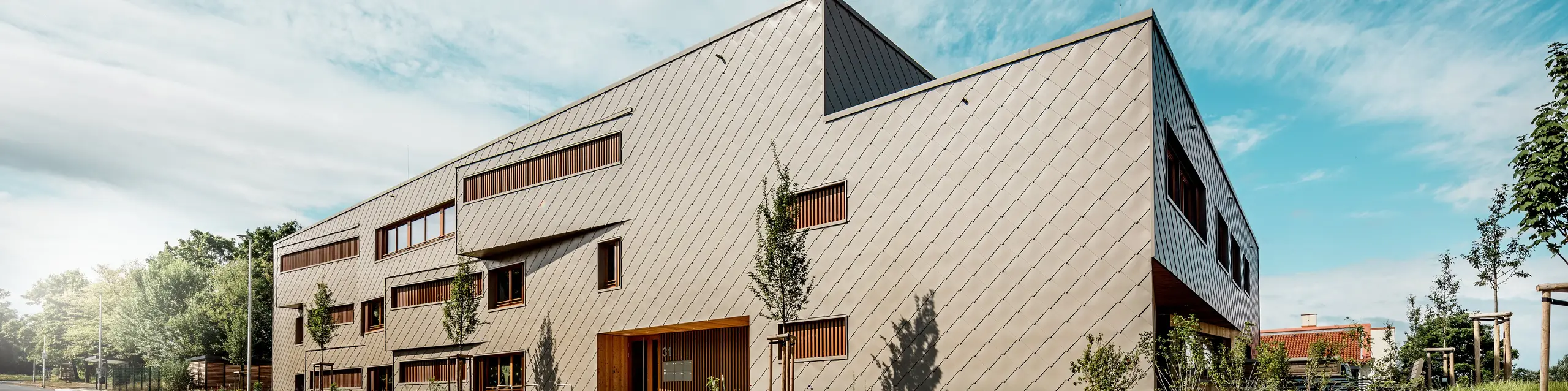 Aus einer markanten Untersichtperspektive präsentiert sich die Kindertagesstätte in Alzenau mit ihrer prägnanten, dynamischen Linienführung und der hochwertigen PREFA Aluminiumfassade in Bronze. Die diagonale Anordnung der Wandrauten 44 × 44 verleiht dem Baukörper eine klare Struktur und eine elegante, homogene Oberfläche. Versetzte Fensteröffnungen und vertikale Holzlamellen setzen rhythmische Akzente, während die reduzierte Formensprache moderne Zurückhaltung signalisiert. Die Kombination aus langlebigem Aluminium, natürlichem Holz und einem durchdachten Grünkonzept entlang der Straße unterstreicht den hohen Anspruch an Nachhaltigkeit und Bauqualität. Dieses Bild zeigt eindrucksvoll, wie funktionale Architektur mit PREFA Produkten gestalterisch überzeugen und städtebaulich Maßstäbe setzen kann.