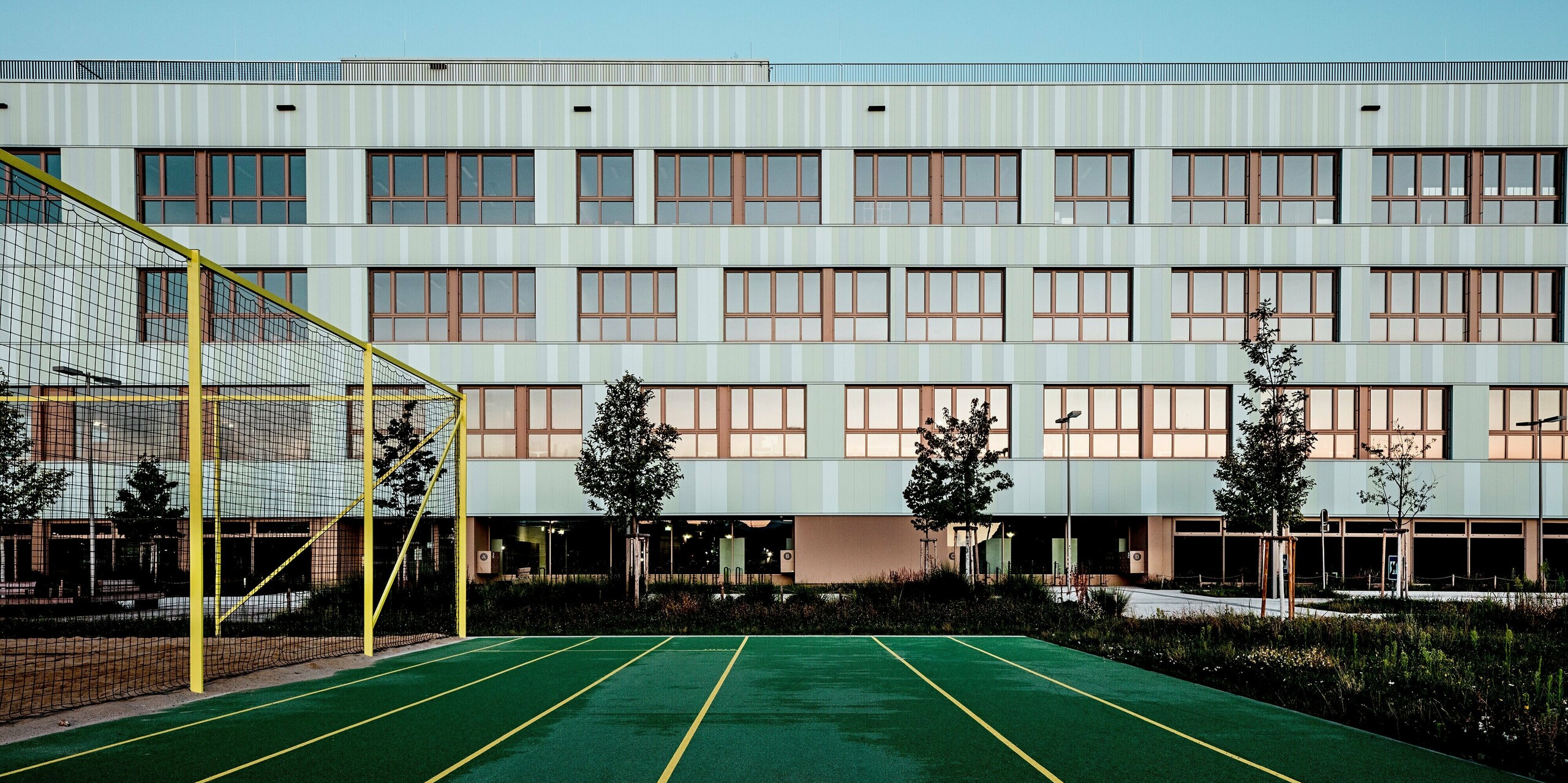 Das Bild zeigt die AHS Reininghaus in Graz mit ihrer modernen und ästhetischen Fassade aus PREFA Sidings in Sonderfarben. Die sanften Grüntöne der Fassade harmonieren perfekt mit den markanten braunen Fensterrahmen, die dem Gebäude einen eleganten Kontrast verleihen. Im Vordergrund ist ein Sportplatz mit einem grünen Laufband und gelben Markierungen zu sehen, was die Nutzung des Gebäudes für Bildung und Sport unterstreicht. Der Platz ist von schön angelegten Grünflächen und Bäumen umgeben, die das moderne Design der Architektur ergänzen. Die Verwendung von PREFA Sidings bietet nicht nur optische Schönheit, sondern auch herausragende Langlebigkeit und Widerstandsfähigkeit gegen Witterungseinflüsse. Die Kombination aus ansprechendem Design und funktionalen Elementen wie der Sporteinrichtung und den gepflegten Außenbereichen macht die AHS Reininghaus zu einem herausragenden Beispiel für zeitgenössische Architektur und nachhaltiges Design.