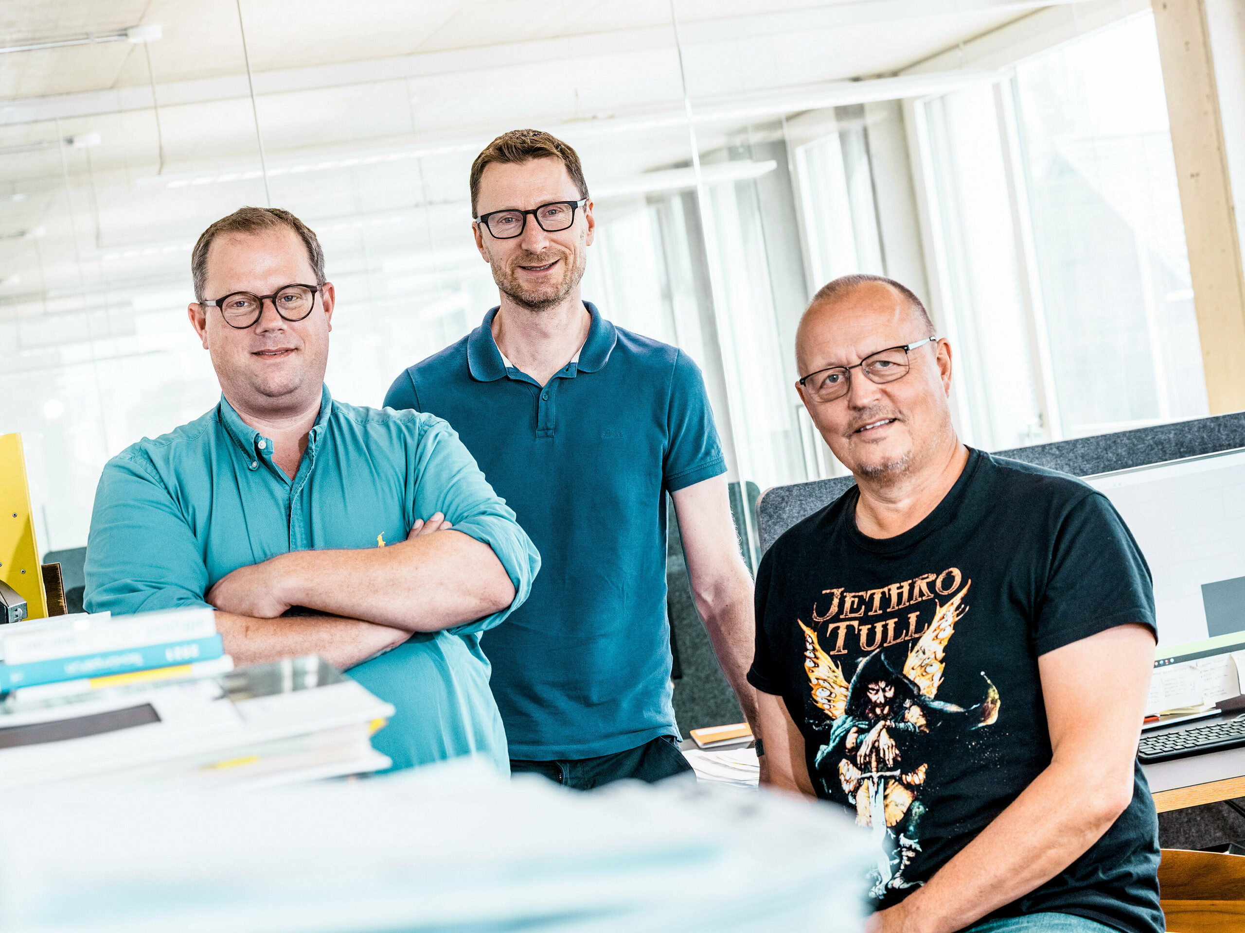 Teamfoto der Kaundbe Architekten in ihrem modernen Büro: Die drei Architekten stehen selbstbewusst vor ihren Arbeitsplätzen und technischen Unterlagen. Im Vordergrund liegen Planstapel und Projektmappen, im Hintergrund sind Computerbildschirme mit Konstruktionszeichnungen zu sehen. Die Szene vermittelt Kompetenz, Zusammenarbeit und technische Präzision – zentrale Werte bei der Planung und Umsetzung von Fassadenlösungen mit PREFA-Aluminiumprodukten. 