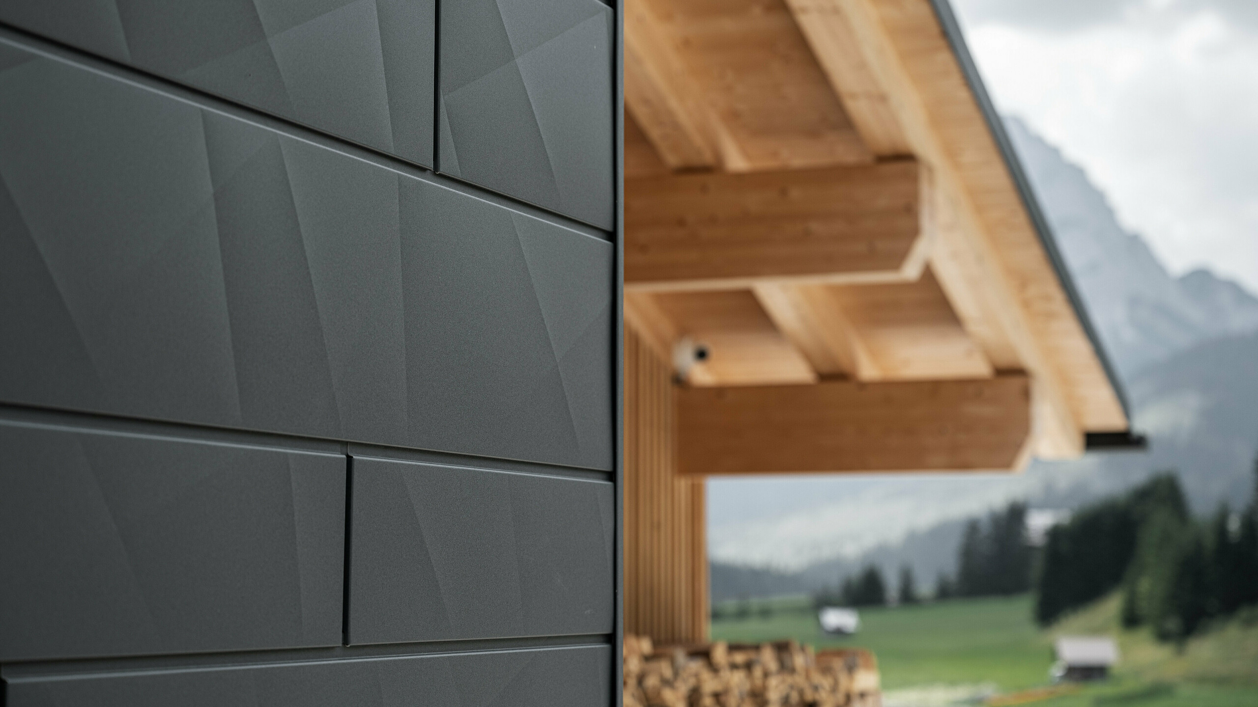 Närbild av Fasadpanel.X i P.10 Svart från PREFA på Casa Hoffe i Sappada, Italien. Fasadsystemet i aluminium är skickligt monterat i en modern, abstrakt design som bildar en tilltalande kontrast till de synliga träelementen i takstolen. Den innovativa fasadlösningen smälter in sömlöst i den alpina miljön och står för en kombination av funktionalitet, hållbarhet och estetisk byggkultur.