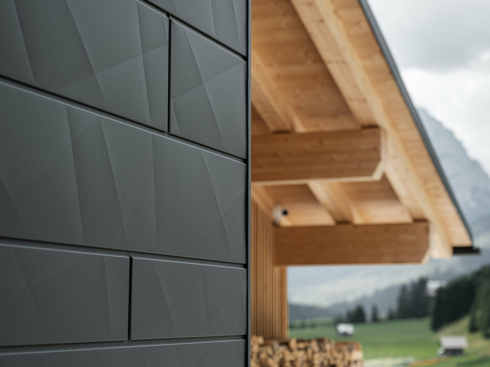 Närbild av Fasadpanel.X i P.10 Svart från PREFA på Casa Hoffe i Sappada, Italien. Fasadsystemet i aluminium är skickligt monterat i en modern, abstrakt design som bildar en tilltalande kontrast till de synliga träelementen i takstolen. Den innovativa fasadlösningen smälter in sömlöst i den alpina miljön och står för en kombination av funktionalitet, hållbarhet och estetisk byggkultur.