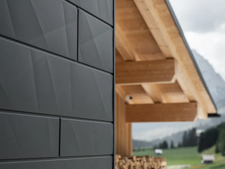 Närbild av Siding.X fasadpanelerna i P.10 svart från PREFA på Casa Hoffe i Sappada, Italien. Fasadsystemet i aluminium är omsorgsfullt monterat i en modern, abstrakt design som bildar en attraktiv kontrast till de synliga träelementen i takstolen. Den innovativa fasadlösningen smälter sömlöst in i den alpina atmosfären och illustrerar kombinationen av funktionalitet, hållbarhet och estetisk byggnadskultur.