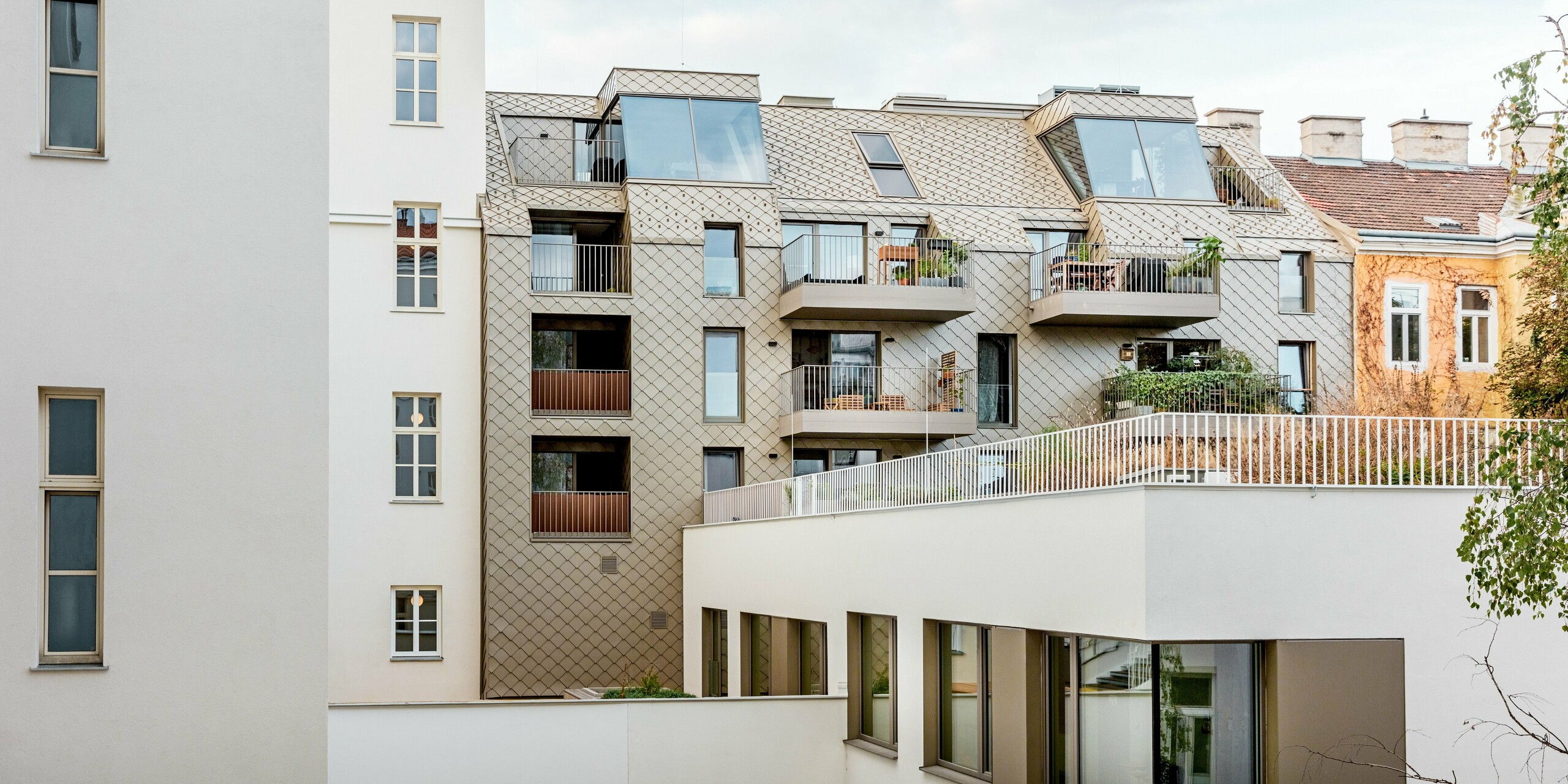 Das Bild zeigt die moderne Erweiterung eines Wohngebäudes in Wien, bei der PREFA Rauten 29×29 in einer warmen Bronzeoptik verwendet wurden. Die Rauten erstrecken sich elegant über das Dach und die Fassade und verleihen dem Gebäude eine zeitgemäße Note, die sich harmonisch in die historische Umgebung einfügt. Besonders auffällig sind die großzügigen Balkone und Fensterflächen, die mit den klaren Linien der Fassade und dem geometrischen Muster der Rauten kontrastieren. Diese gelungene Kombination aus Tradition und Moderne sorgt für eine visuelle Spannung und hebt die Architektur des Objekts hervor.