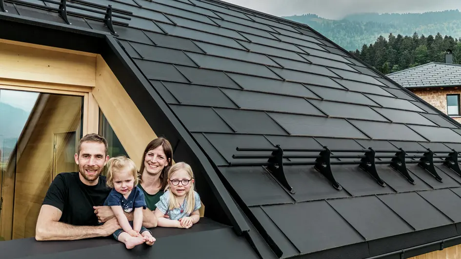 De två byggherrarna njuter tillsammans med sina barn av utsikten från loggian i sitt nybyggda hus, som är utrustat med eleganta PREFA-solar takplattor i P.10 svart.