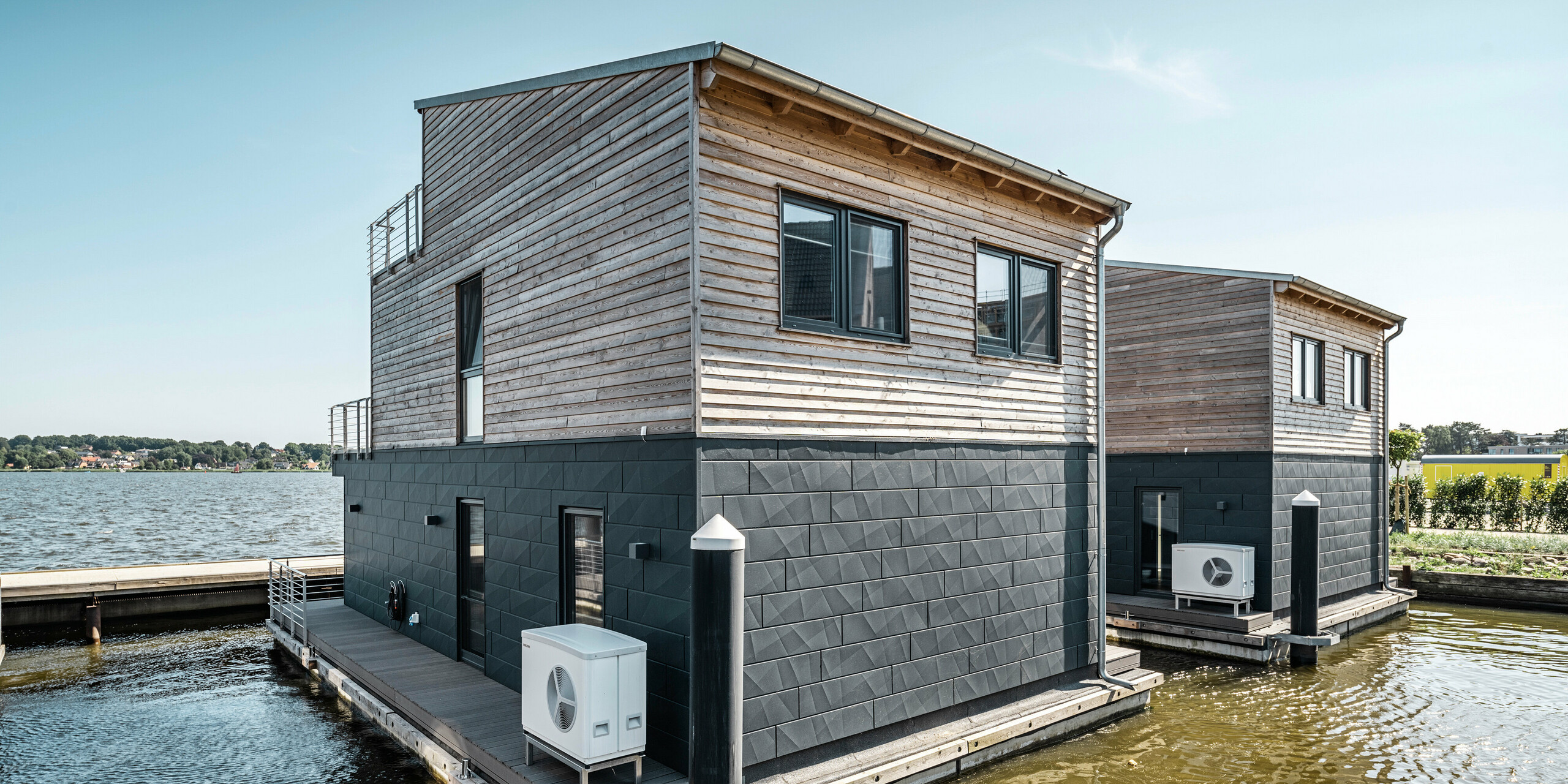 Zwei nebeneinander liegende Wasserhäuser im Pionierhafen Schleswig, stilvoll kombiniert mit PREFA Siding.X in P.10 Anthrazit und natürlichem Holz im oberen Bereich, fügen sich nahtlos in die Uferlandschaft ein. Die Häuser stehen auf stabilen Schwimmkörpern und bieten durch ihre großen Fensterfronten und Außenterrassen eine offene und luftige Wohnatmosphäre. Die Anlage zeichnet sich durch ihre nachhaltige Bauweise und die Integration moderner Haustechnik, wie die außen montierte Wärmepumpe, aus, die eine umweltbewusste Lebensweise in exklusiver Lage am Wasser ermöglicht.