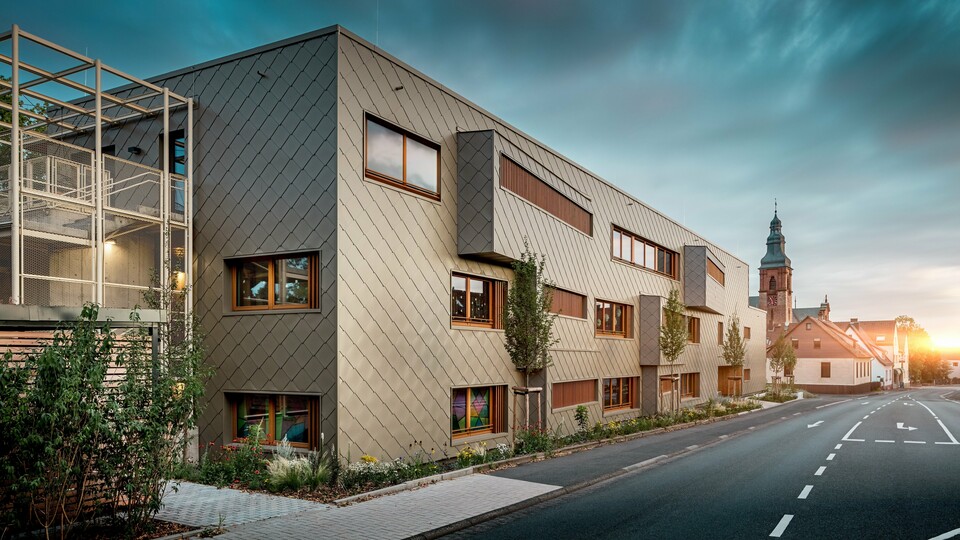 Im sanften Licht des Sonnenuntergangs zeigt sich die Kindertagesstätte in Alzenau von ihrer stimmungsvollsten Seite. Die Fassade aus bronzefarbenen PREFA Wandrauten 44 × 44 reflektiert das Abendlicht und betont die hochwertige, strukturierte Oberfläche des modernen Hybridgebäudes. Die klar gegliederte Architektur mit versetzten Fensterbändern und vertikalen Holzlamellen steht in spannendem Kontrast zur historischen Kirche im Hintergrund. Links im Bild sorgt ein filigranes Metallgerüst für leichte Transparenz beim Außentreppenhaus. Der gepflegte Grünstreifen entlang des Gebäudes unterstreicht den ökologischen Anspruch. Dieses Bild verbindet eindrucksvoll moderne Baukultur, nachhaltige PREFA Aluminiumtechnologie und harmonische Einbettung in das historische Stadtbild von Alzenau.