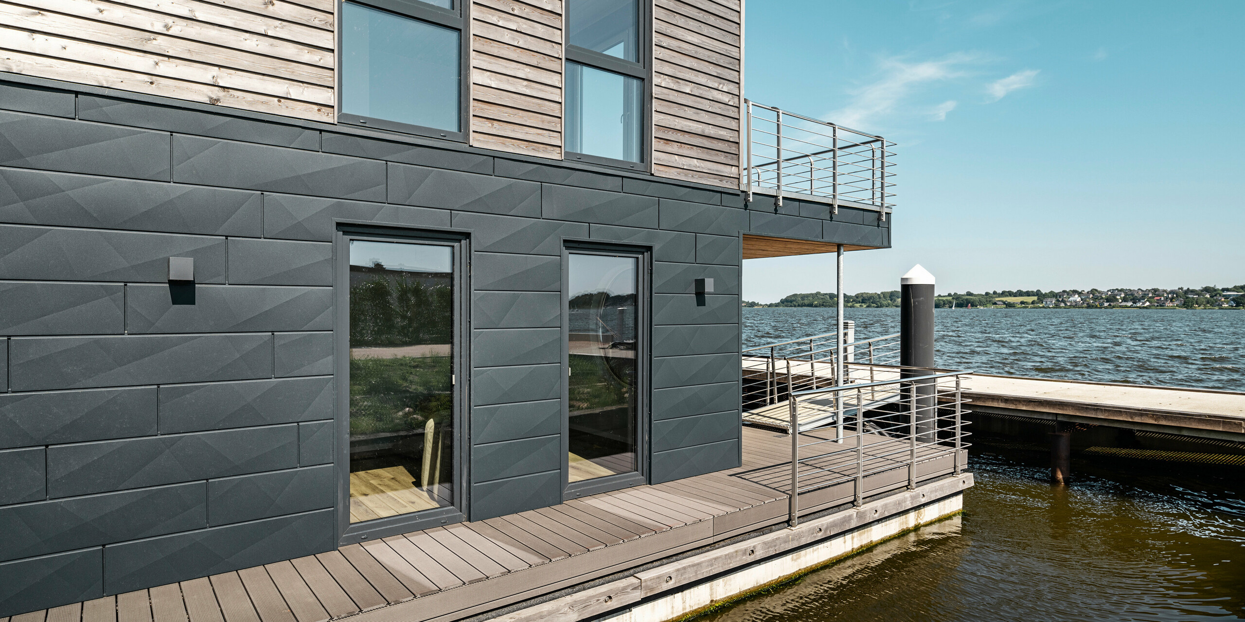 Seitliche Ansicht eines schwimmenden Hauses im Pionierhafen Schleswig, das mit robusten PREFA Siding.X in P.10 Anthrazit verkleidet ist, vor dem Hintergrund der breiten Schlei. Das Haus präsentiert stolz die Kombination aus nachhaltigem Holz und dem beständigen Aluminium an der Fassade, die neben ihrer Funktionalität auch eine stilvolle Eleganz ausstrahlt. Die großflächigen Fenster bieten eine klare Sicht und verbinden das Innere mit der malerischen Wasserlandschaft.