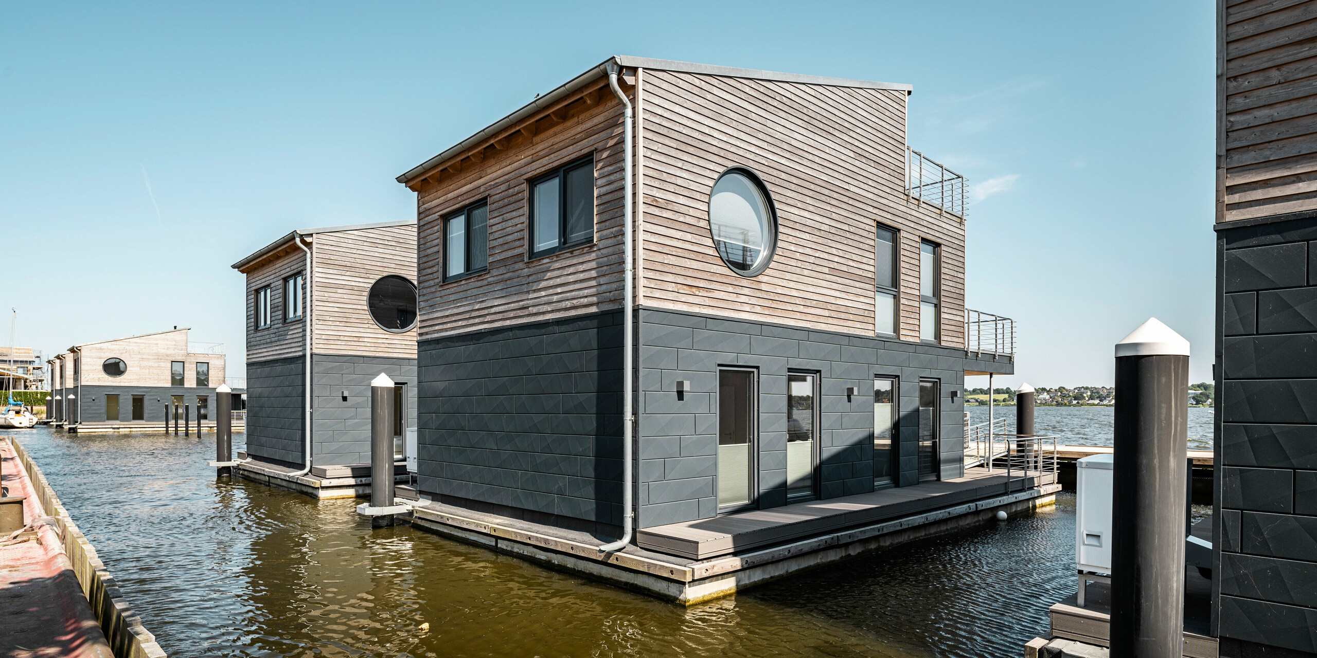Perspektivische Ansicht einer Reihe von schwimmenden Häusern im Pionierhafen Schleswig, deren Fassaden mit PREFA Siding.X in P.10 Anthrazit für eine starke Witterungsbeständigkeit gestaltet sind. Die runden Fenster ergänzen das moderne Design und bieten einen maritimen Touch, während die Holzelemente der oberen Geschosse eine natürliche Wärme ausstrahlen. Die Konstruktionen sind fest verankert in der ruhigen Wasserstraße, was den Bewohnern einen unmittelbaren Zugang zum Wasser und eine einzigartige Wohnatmosphäre ermöglicht.