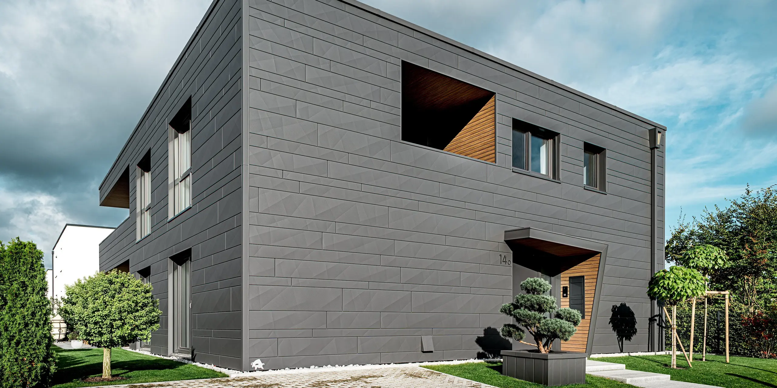Das moderne Einfamilienhaus in Heidenheim zeigt eine vollständig mit PREFA Siding.X in P.10 Dunkelgrau verkleidete Aluminiumfassade, deren markante Kantungen und unterschiedlichen Paneelbreiten dem klaren Baukörper eine dynamische Struktur verleihen. Der holzverkleidete Eingangsbereich bildet einen warmen Kontrast zur eleganten Metalloberfläche. Im Vordergrund steht eine sorgfältig gestaltete Gartenfläche. Die robuste PREFA Aluminiumfassade und das integrierte Quadratrohr bieten langlebigen Witterungsschutz, hohe Stabilität und ein zeitlos modernes Erscheinungsbild.