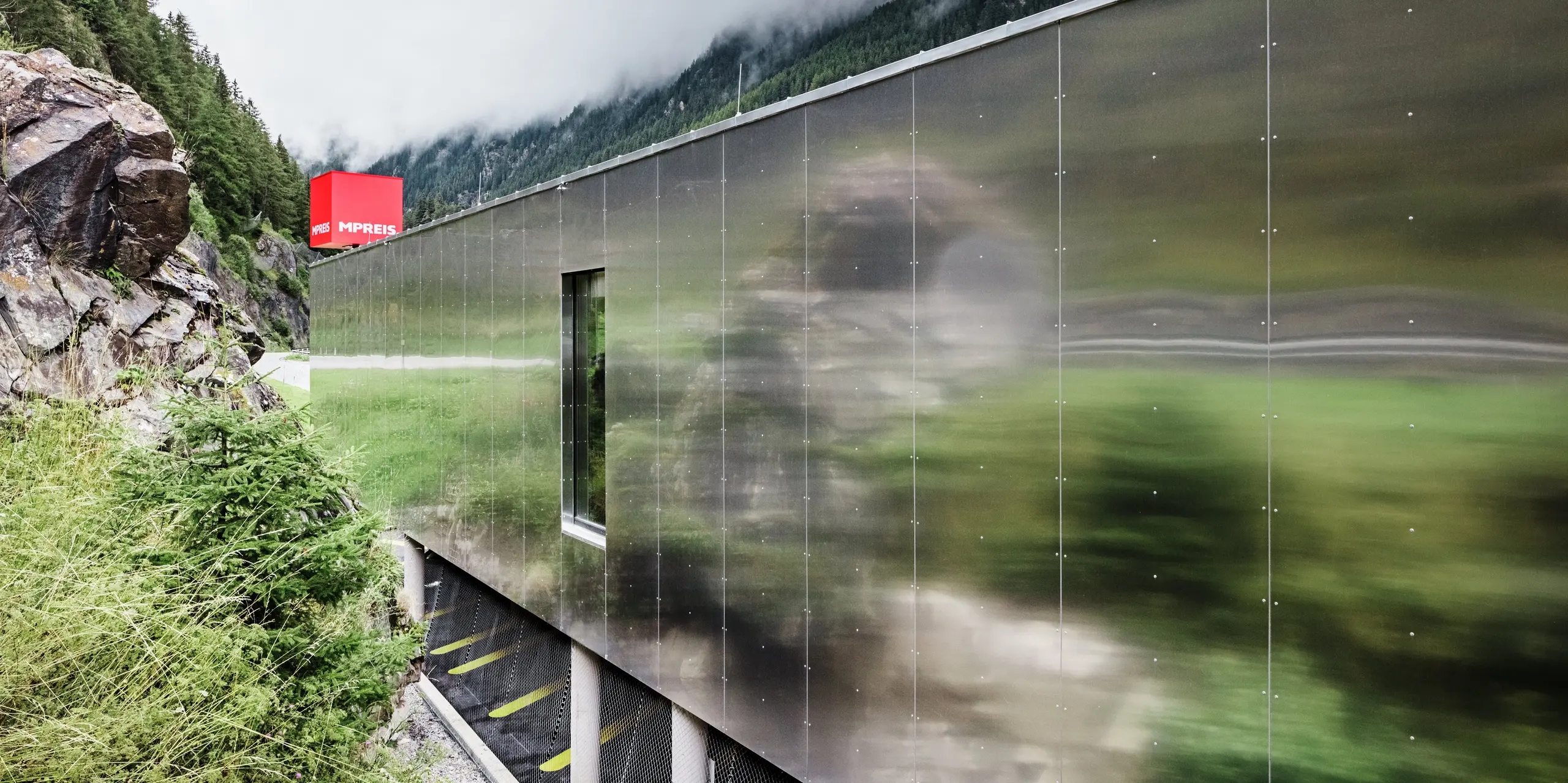 Nahaufnahme der verspiegelten Fassade des MPREIS Supermarktes in Sölden, Tirol. Die Aluminium Verbundplatten in der Sonderfarbe Reflective Natural spiegeln die umgebende Landschaft der Ötztaler Alpen wider. Die fugenlos montierten Platten erzeugen eine mattglänzende Oberfläche, die moderne Architektur mit der Natur verschmelzen lässt.