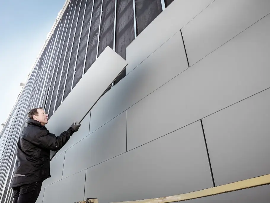 Foton av en delvis färdigställd fasad som är beklädd med PREFA Sidings i bredden 500. Plåtslagaren monterar ytterligare en fasadpanel. Bilden är tagen underifrån. 