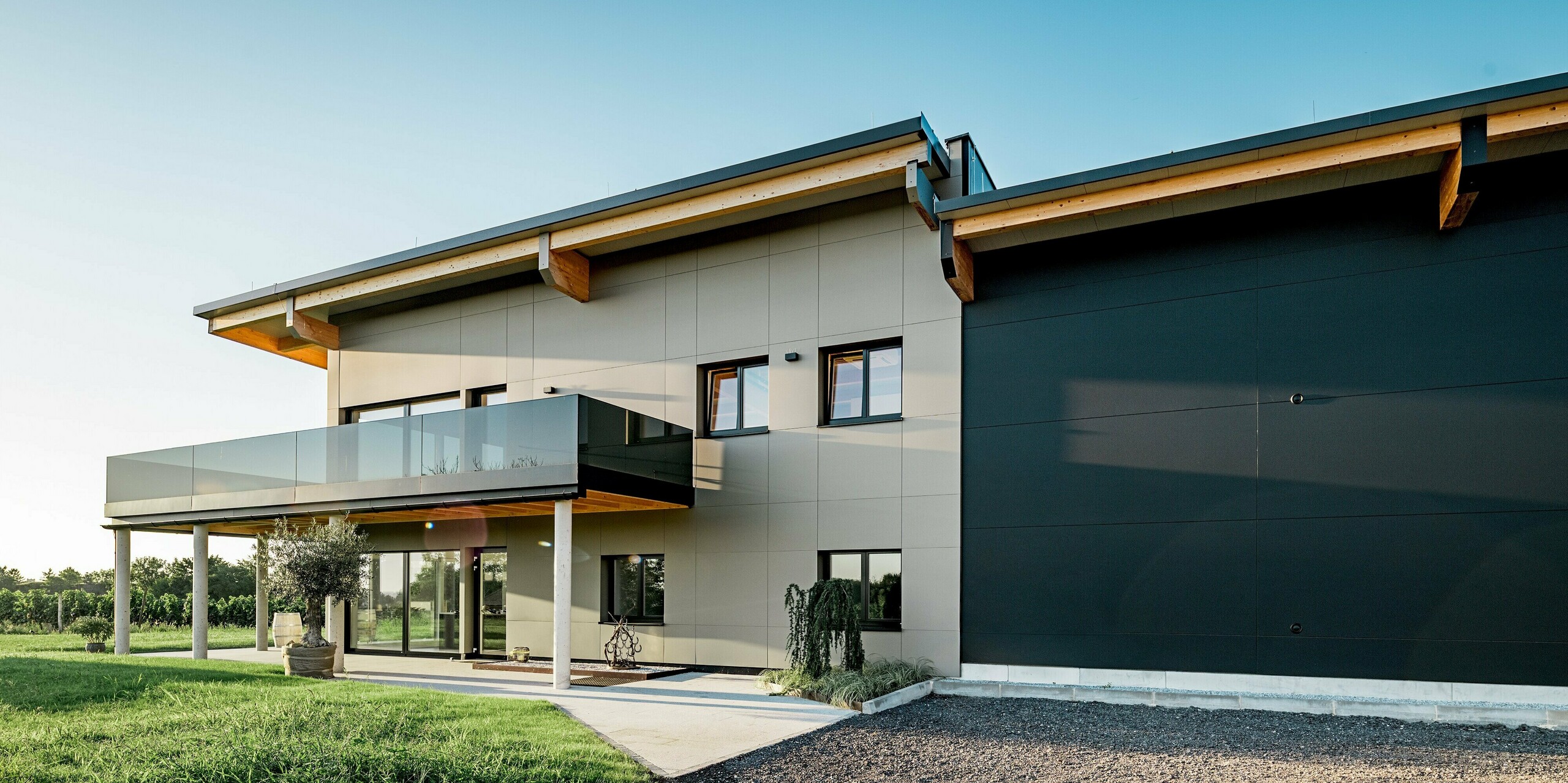 Seitenansicht des Weinguts Nigl in Perchtoldsdorf mit kontrastreicher PREFA Aluminiumfassade in den Farben P.10 Prefabronze und P.10 Schwarz. Links dominiert die helle Fassadenseite mit großzügigem Balkon samt Glasbrüstung und filigranen Stützen, während rechts die tiefschwarze Aluminiumfläche mit klaren Linien Ruhe und Stärke ausstrahlt. Sichtbare Holzdachträger unterstreichen den modernen Landhausstil. Die Kombination aus hochwertigen PREFA Verbundplatten, Glas, Holz und Beton schafft eine zeitlose, elegante Architektur mit maximaler Witterungsbeständigkeit und Langlebigkeit – perfekt für repräsentative Bauten im Weinbauumfeld.