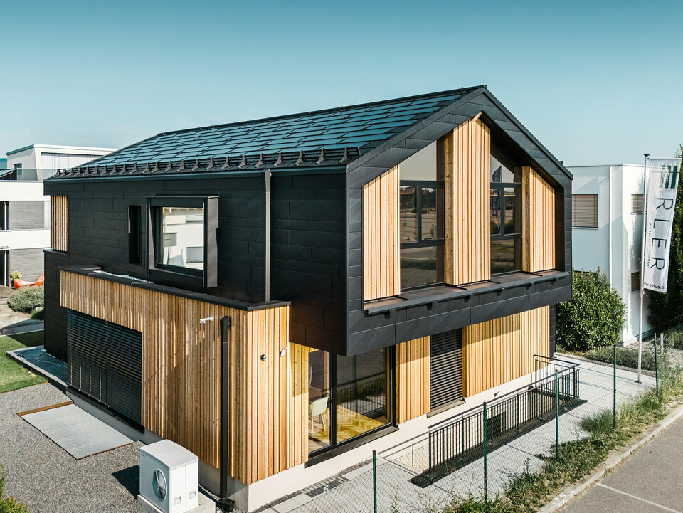 Framsidan av modellhuset Autark i prefabricerade huscentret Blaue Lagune, Vösendorf (AT). Taket är täckt med svarta PREFA soltakplattor och takplatta R.16 med högkvalitativ P.10-beläggning. Fasaden består av en kombination av vertikala träelement och PREFA Fasadpanel.X i P.10 svart, vilket ger byggnaden en modern och hållbar karaktär. I höger bildkant vajar Erler Bau GmbH:s flagga.