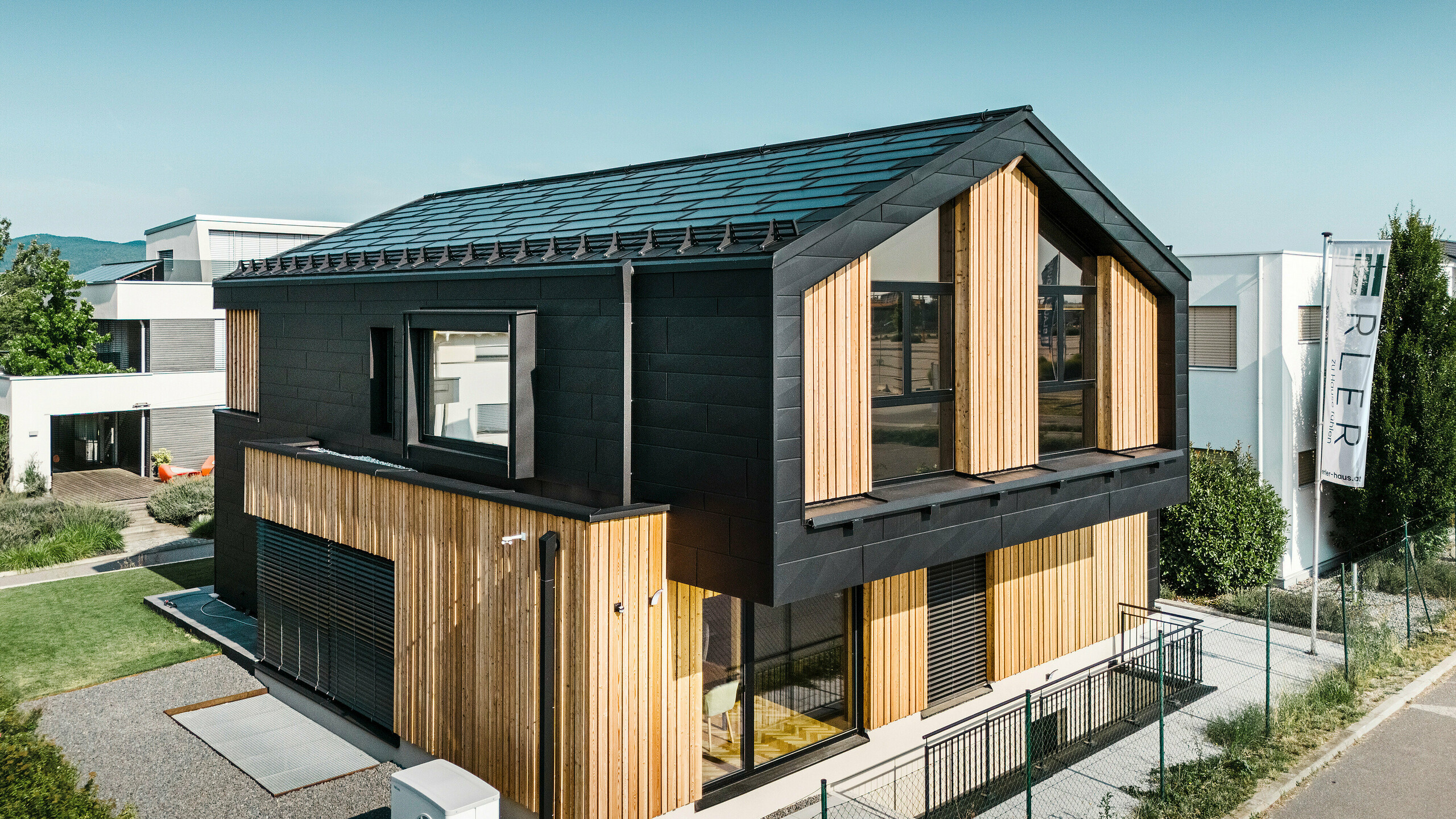 Framsidan av modellhuset Autark i prefabricerade huscentret Blaue Lagune, Vösendorf (AT). Taket är täckt med svarta PREFA soltakplattor och takplatta R.16 med högkvalitativ P.10-beläggning. Fasaden består av en kombination av vertikala träelement och PREFA Fasadpanel.X i P.10 svart, vilket ger byggnaden en modern och hållbar karaktär. I höger bildkant vajar Erler Bau GmbH:s flagga.