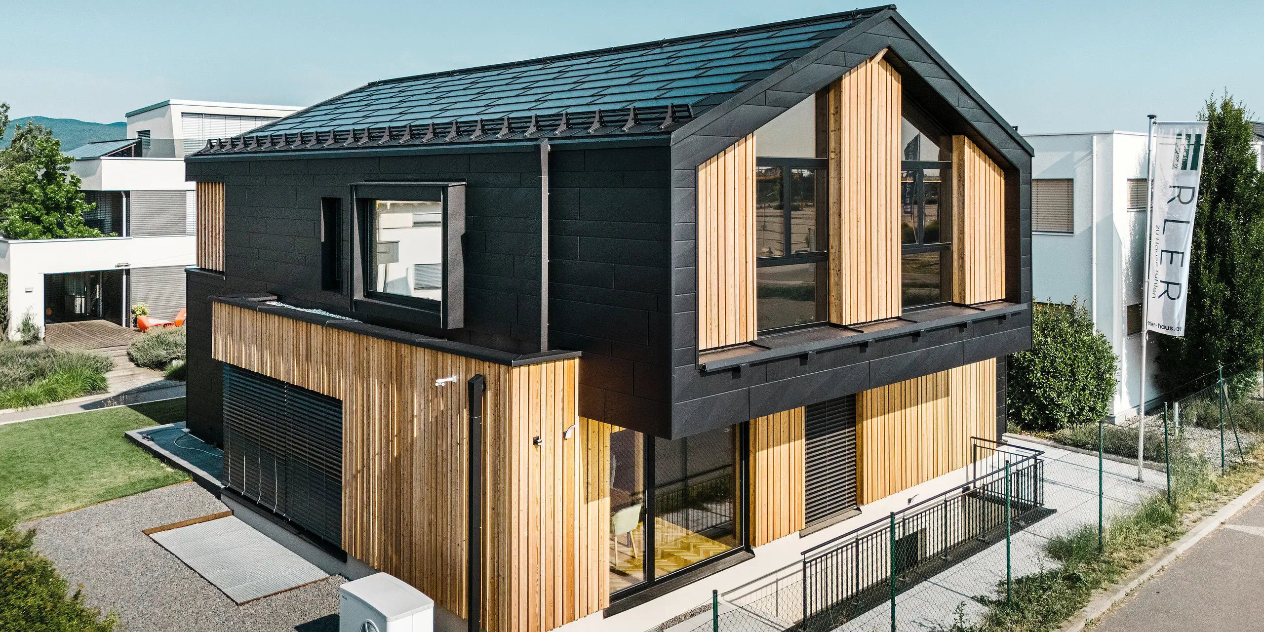 Framsidan av modellhuset Autark i prefabricerade huscentret Blaue Lagune, Vösendorf (AT). Taket är täckt med svarta PREFA soltakplattor och takplatta R.16 med högkvalitativ P.10-beläggning. Fasaden består av en kombination av vertikala träelement och PREFA Fasadpanel.X i P.10 svart, vilket ger byggnaden en modern och hållbar karaktär. I höger bildkant vajar Erler Bau GmbH:s flagga.