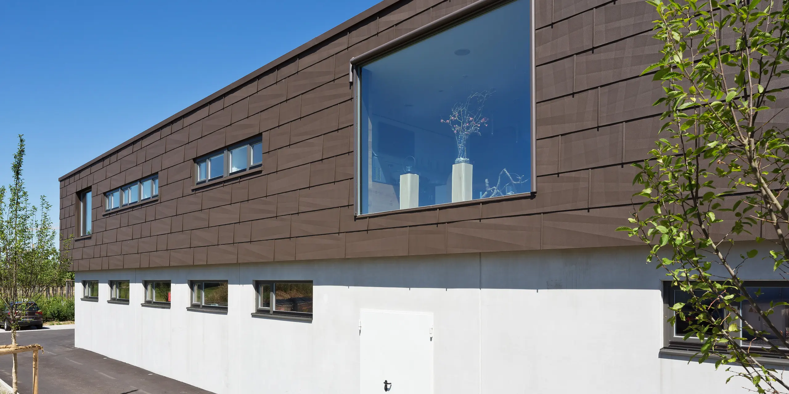 Die Seitenansicht des Coffeeteam-Firmengebäudes in München beeindruckt mit einer großflächigen PREFA Fassade aus FX.12 Fassadenpaneelen in P.10 Braun. Das markante Fassadenbild lebt von den charakteristischen Kantungen, die spannende Licht- und Schatteneffekte erzeugen. Ein großzügiges Panoramafenster setzt einen architektonischen Akzent, während die langlebige, witterungsbeständige und nahezu wartungsfreie PREFA Aluminiumfassade moderne Gestaltung und Funktionalität vereint.