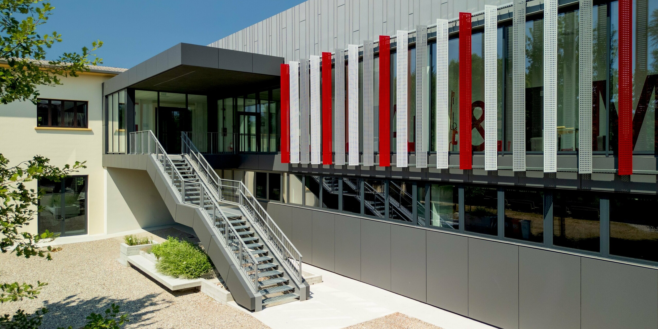 Einladender Innenhofbereich der Scuola Arte e Mestieri F. Bertazzoni in Suzzara mit einer eleganten Außentreppe, die zum verglasten Obergeschoss führt. Die zweigeschossige PREFA Aluminiumfassade in Dunkelgrau besticht durch ihr lebendiges Wechselspiel aus perforierten Elementen in Hellgrau und feurig roten Fassadenstreifen, hinter denen sich großformatige Lettern abzeichnen. PREFALZ und PREFABOND von PREFA überzeugen mit ihrer präzisen Verarbeitung, dauerhaften Oberflächenqualität und hervorragenden Witterungsbeständigkeit und verleihen dem Schulgebäude eine unverwechselbare Identität mit kraftvollem gestalterischen Ausdruck.