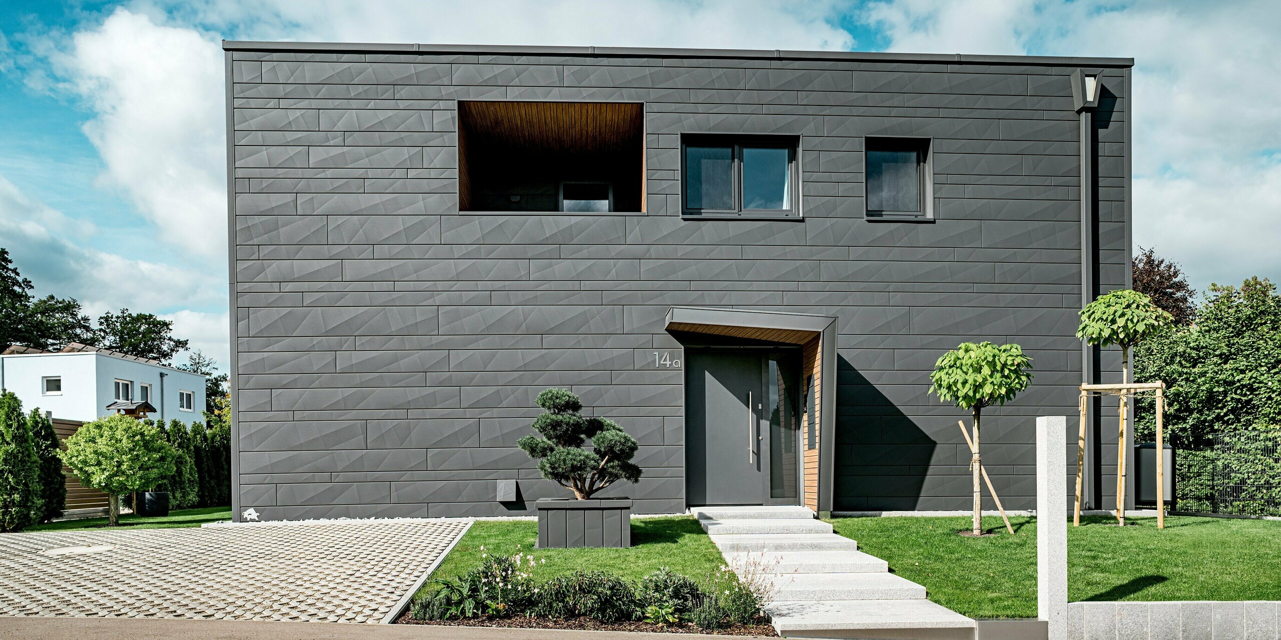 Frontalansicht eines modernen Einfamilienhauses in Heidenheim mit durchgehender PREFA Siding.X Fassade in P.10 Dunkelgrau, deren markante Linien und unterschiedlichen Paneelbreiten ein lebendiges Fassadenbild erzeugen. Der holzverkleidete Eingangsbereich und die darüberliegende Loggia setzen warme Akzente zur klaren Aluminiumarchitektur. Im Vordergrund strukturieren formgeschnittene Gehölze, junger Baumbestand und ein gepflasterter Vorplatz die Szene. Die robuste PREFA Aluminiumoberfläche bietet langlebigen Witterungsschutz und unterstreicht das moderne, präzise Erscheinungsbild des Gebäudes.