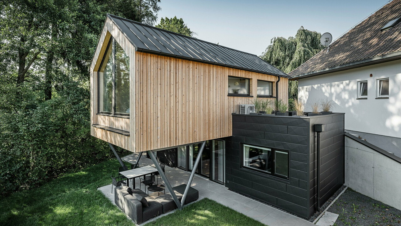 Das 'Woodstock Chalet 5' beeindruckt mit einer eindrucksvollen Kombination aus natürlichen Holzoberflächen und den modernen PREFA Siding.X Fassadenelementen in Anthrazit. Die überhängende Holzstruktur des Obergeschosses ragt markant über die dunklen Fassadenelemente, die das Erdgeschoss verkleiden, und erzeugt so ein Spiel aus Licht und Schatten. Die klaren Linien und die robuste Konstruktion der Siding.X Elemente ergänzen das innovative Design des Gebäudes und bieten einen spannenden visuellen Kontrast zur umgebenden Vegetation und dem traditionellen Gebäude im Hintergrund. Am Dach des Chalets wurde das PREFALZ Dachsystem in Form einer gleichmäßigen Stehfalzdeckung verlegt. Am rechten Rand des Erdgeschosses ist das PREFA Dachentwässerungssystem zu erkennen, das analog zur Aluminiumfassade in Anthrazit gehalten ist. Für die Dachentwässerung wurden der Wasserfangkasten gefolgt vom Ablaufrohr montiert.