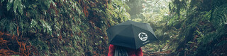Kvinna vandrar i röd jacka med PREFA-paraply och gymnastikpåse – symboliserar PREFA:s arbete med miljöskydd, hållbarhet, kretslopp och återvinning