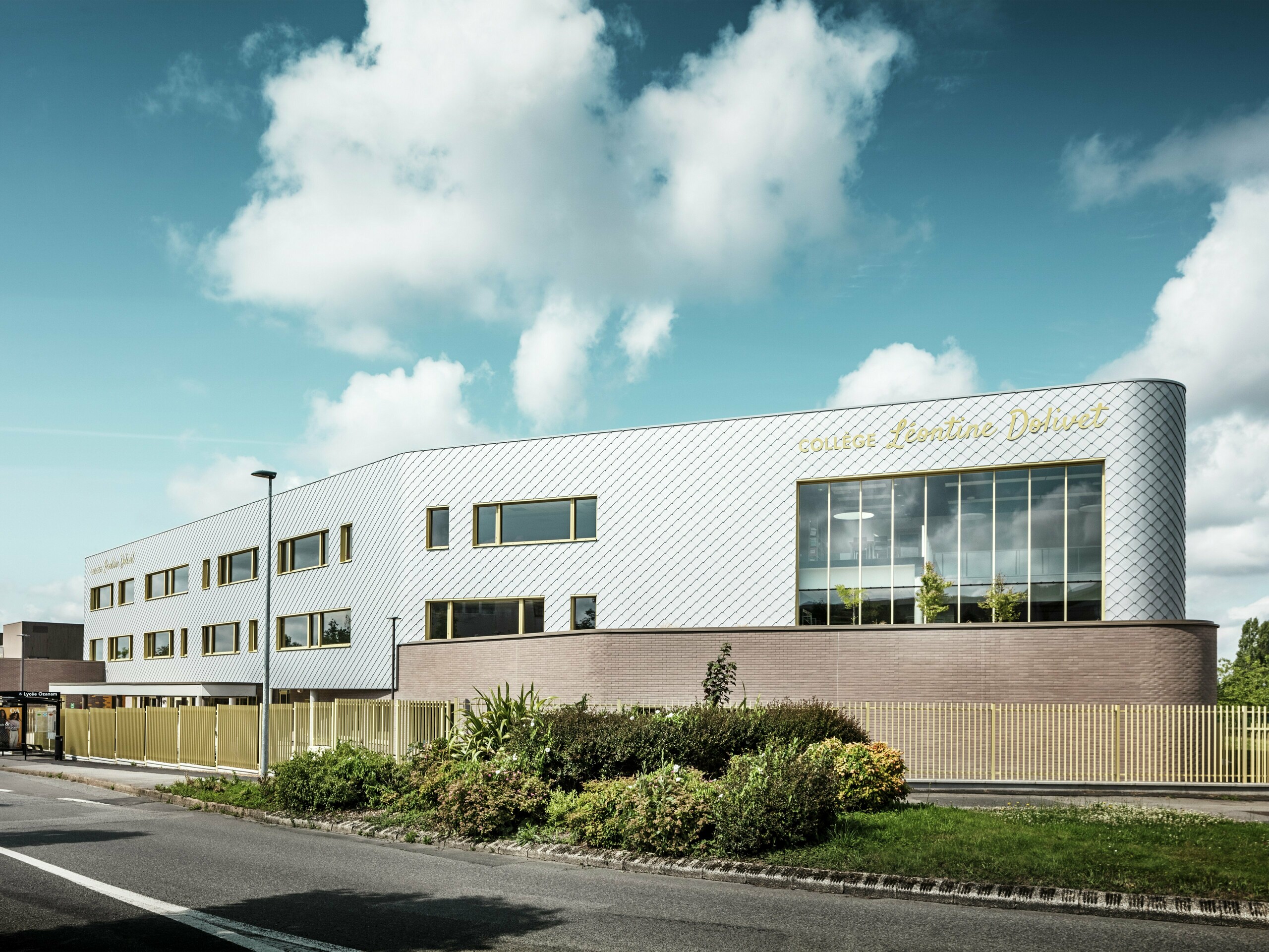 Ansicht des Collège Léontine Dolivet mit markanter, silbrig glänzender Aluminiumfassade aus PREFA Wandrauten 29 × 29 in Silbermetallic. Die Fassade wirkt dynamisch durch das diagonale Rautenmuster, während große Glasflächen und goldenen Fensterläden spannende Kontraste setzen. Im Vordergrund sind gepflegte Grünflächen und ein moderner Zaun zu sehen, die das Gebäude einrahmen. Die langlebige PREFA Fassadenlösung kombiniert Eleganz, Witterungsbeständigkeit und Nachhaltigkeit – ein architektonischer Eyecatcher in Cesson-Sévigné nahe Rennes.