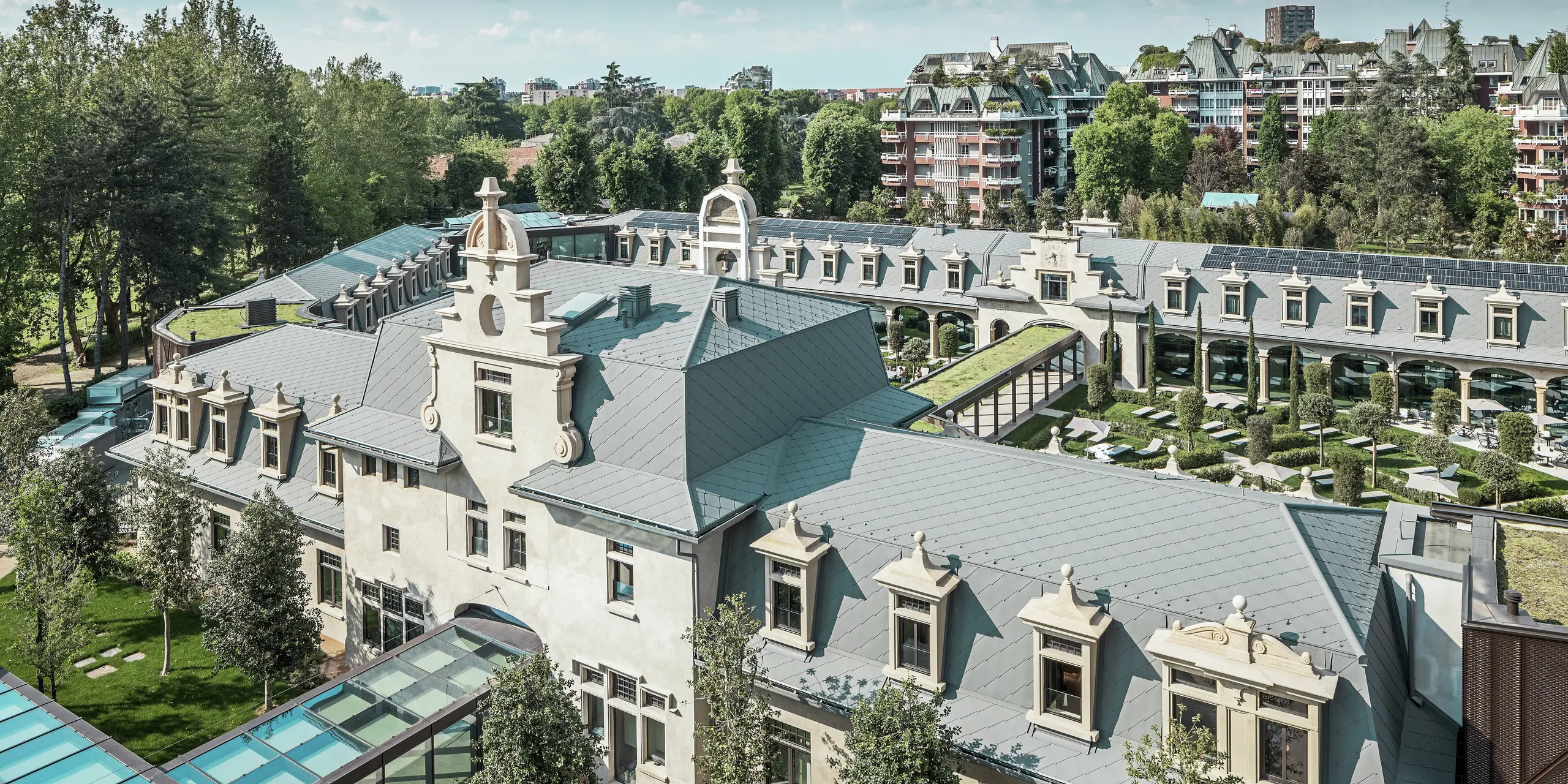 Helhetsbild av den imponerande anläggningen ”De Montel – Terme Milano” med kulturminnesmärkt jugendarkitektur och modern wellnessmiljö. Den stora takbeläggningen med PREFA takromb 44 × 44 i P.10 ljusgrå ger anläggningen en elegant enhetlighet. Integrerade solcellsmoduler, gröna tak och glasförbindelser mellan byggnaderna speglar kraven på hållbarhet och modern design i en urban kontext.