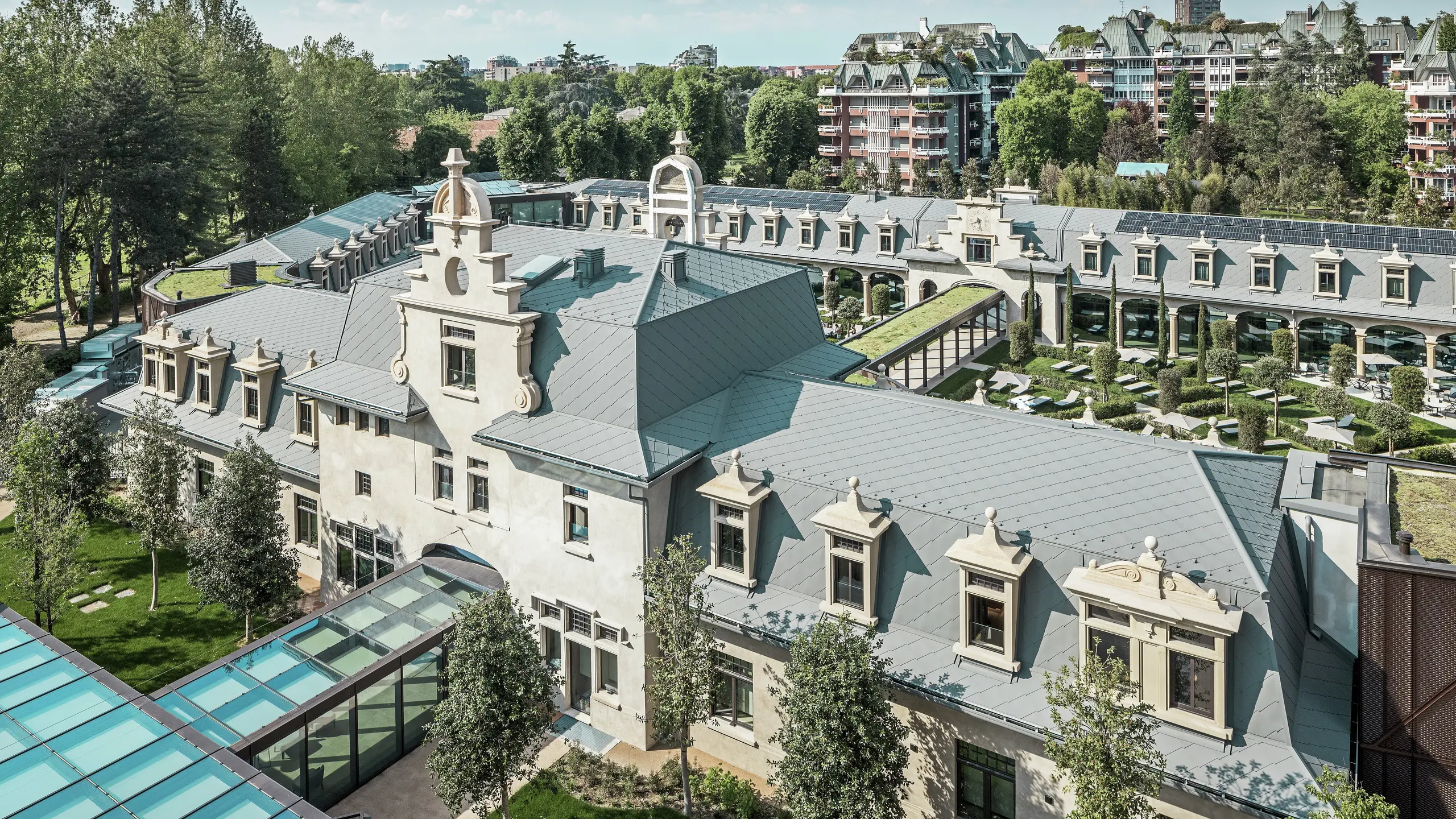 Helhetsbild av den imponerande anläggningen ”De Montel – Terme Milano” med kulturminnesmärkt jugendarkitektur och modern wellnessmiljö. Den stora takbeläggningen med PREFA takromb 44 × 44 i P.10 ljusgrå ger anläggningen en elegant enhetlighet. Integrerade solcellsmoduler, gröna tak och glasförbindelser mellan byggnaderna speglar kraven på hållbarhet och modern design i en urban kontext.