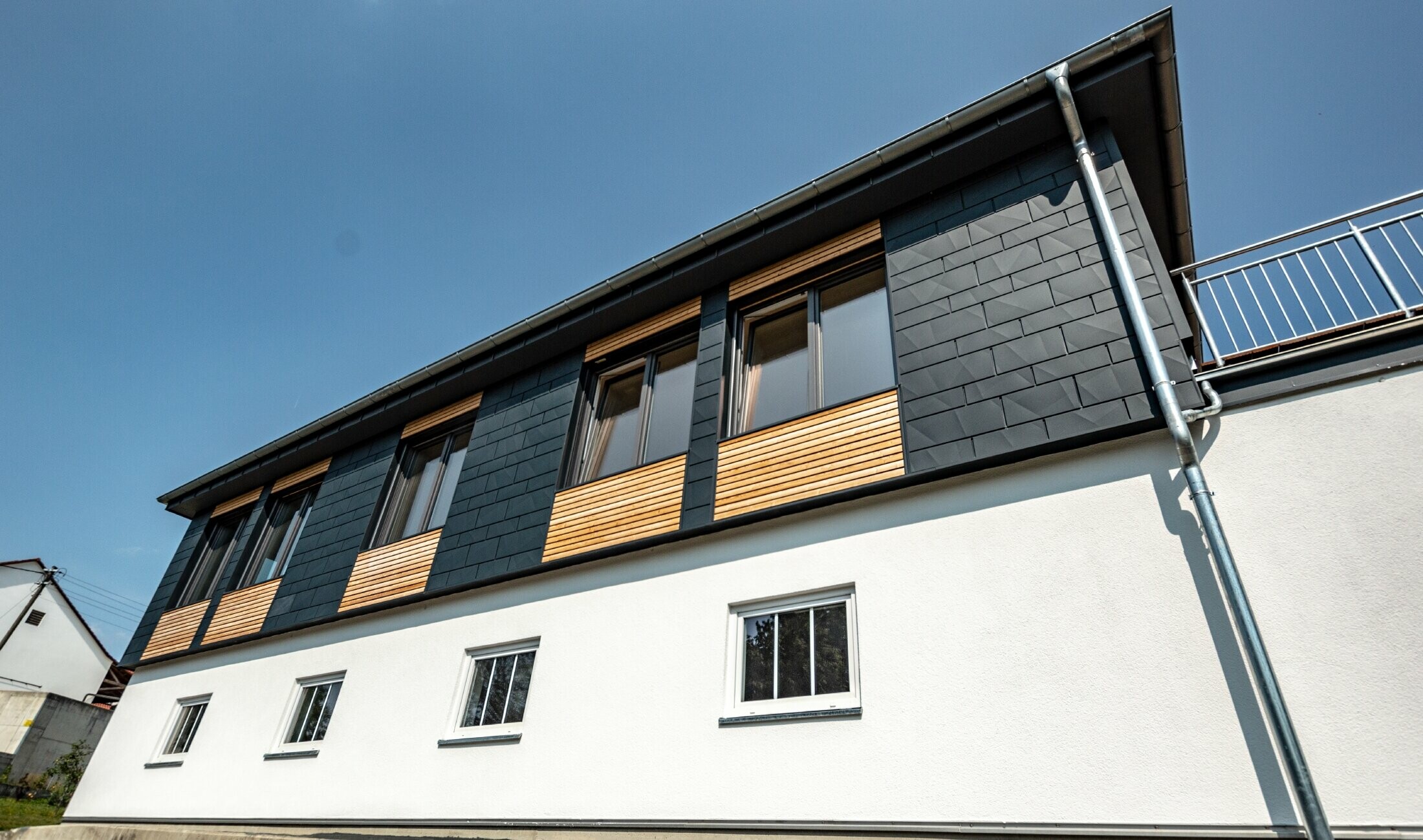 Fasaddesign med olika byggmaterial, aluminium PREFA Siding.X i antracit, en horisontell träfasad och en vit putsfasad.