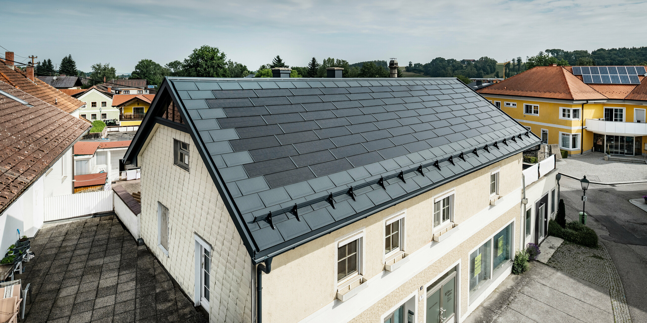 Vy från ovan över ett fristående hus i Mettmach, Österrike. Den äldre byggnaden har renoverats med antracitfärgade PREFA solar takplatta. Snöskyddssystemen och de klara linjerna på solcellstakpanelerna understryker det rena utförandet av PREFAs aluminiumprodukter. Det orangefärgade tegeltaket på grannbyggnaden kontrasterar i bakgrunden. Solcellsmodulerna är installerade på taket till grannhuset i en klassisk on roof-lösning, vilket visar på mångfalden av energieffektiv takdesign.