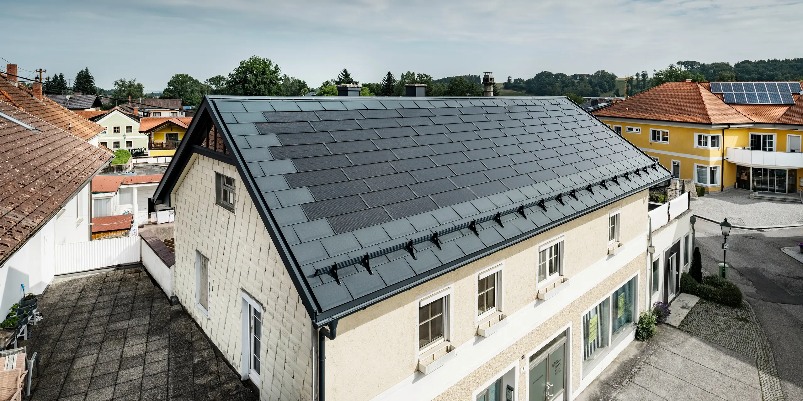 Vy från ovan över ett fristående hus i Mettmach, Österrike. Den äldre byggnaden har renoverats med antracitfärgade PREFA solar takplatta. Snöskyddssystemen och de klara linjerna på solcellstakpanelerna understryker det rena utförandet av PREFAs aluminiumprodukter. Det orangefärgade tegeltaket på grannbyggnaden kontrasterar i bakgrunden. Solcellsmodulerna är installerade på taket till grannhuset i en klassisk on roof-lösning, vilket visar på mångfalden av energieffektiv takdesign.