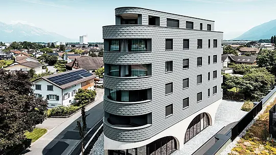 Luftaufnahme des Bernerhauses in Buchs mit markanter, dreieckiger Kubatur und weich gerundeten Balkonen. Die Fassade aus PREFA Wandschindeln in P.10 Hellgrau umschließt das Gebäude wie eine schuppenartige Aluminiumhaut und sorgt für ein lebendiges Spiel aus Licht und Schatten. Über dem massiven Betonsockel mit eleganten Arkaden wirkt die leichte, witterungsbeständige PREFA Aluminiumfassade besonders präzise, langlebig und wartungsarm – ein moderner Eyecatcher inmitten der alpinen Stadtlandschaft.