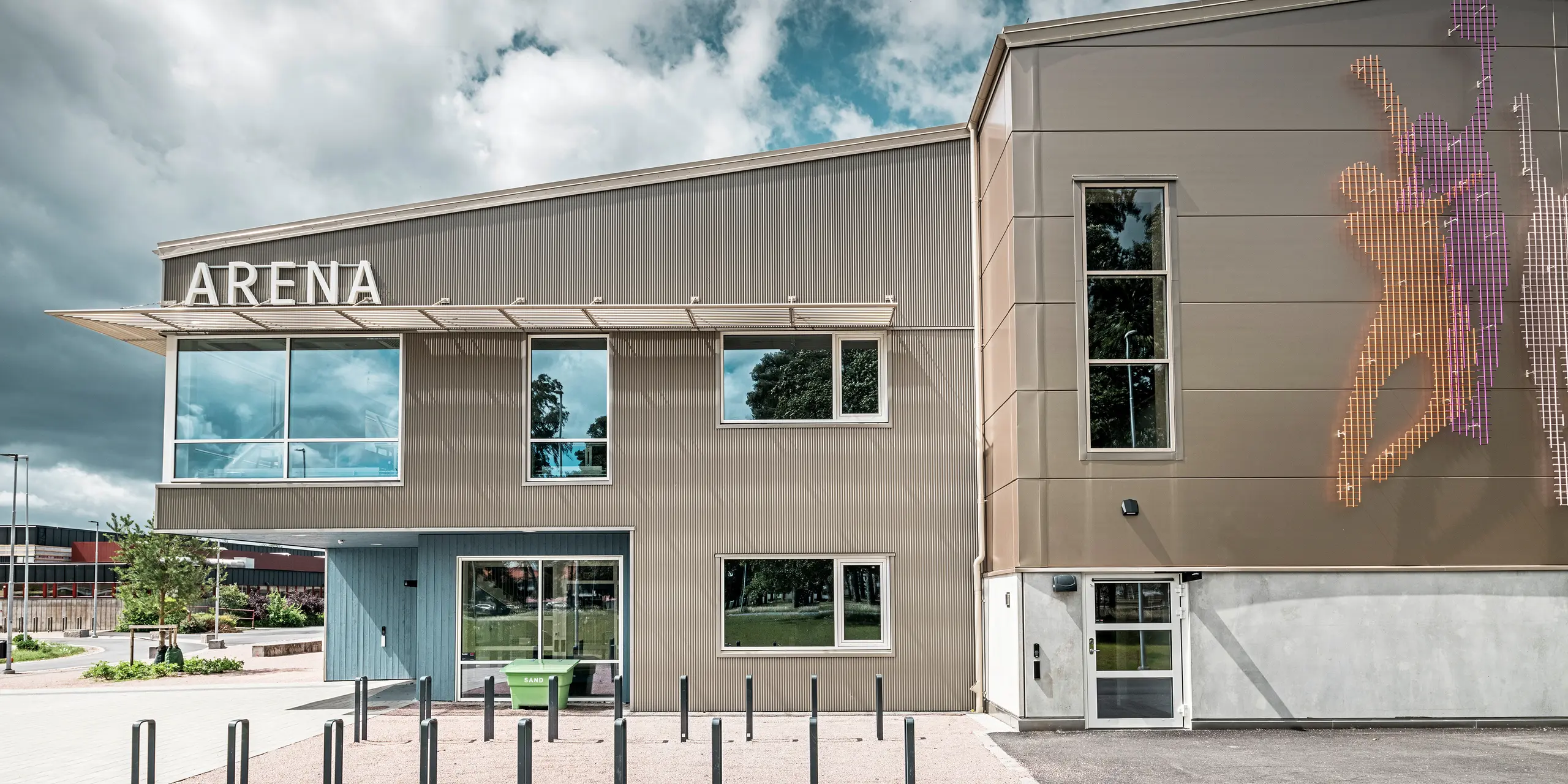 Seitenansicht des Eingangsbereichs zur Park Arena in Värnamo, Schweden, mit moderner PREFA Fassade aus dem Zackenprofil in Bronze. Die Aluminiumfassade ist langlebig und sorgt für markante Licht- und Schatteneffekte. Rechts im Bild befindet sich ein künstlerisch gestaltetes Wandbild, das sportliche Figuren darstellt und die dynamische Atmosphäre der Sporthalle unterstreicht. Die großen Fensterflächen lassen viel Licht in das Gebäude und betonen das zeitgemäße Design.