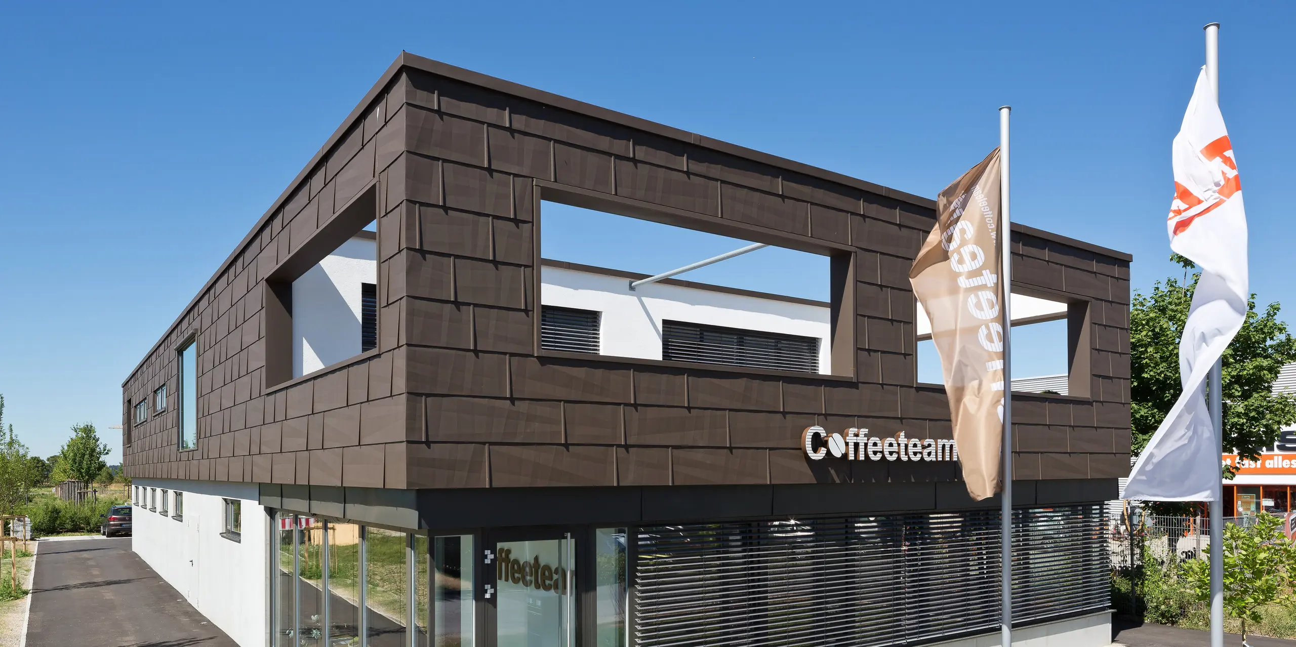 Modernes Firmengebäude von Coffeeteam in München mit markanter Fassadenverkleidung. PREFA Fassadenpaneele FX.12 in P.10 Braun zieren das Bauwerk. Die kantigen Aluminium-Fassadenpaneele erzeugen faszinierende Licht- und Schatteneffekte und betonen die klare Architektur des Gebäudes. Großzügige Fensterflächen und die langlebige, witterungsbeständige sowie wartungsarme PREFA Aluminiumfassade schaffen einen ausdrucksstarken und zeitgemäßen Unternehmensauftritt.