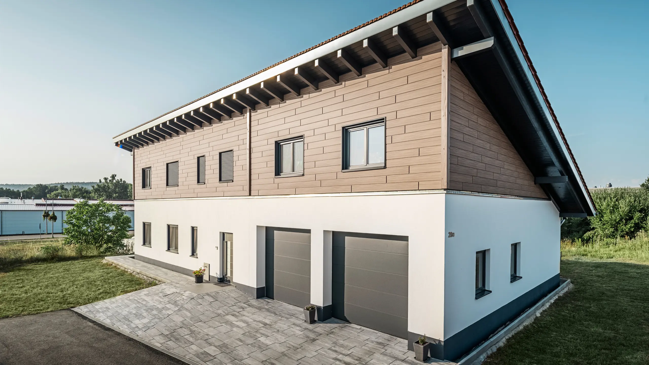 Vy från sidan av en villa med pulpettak i Bruck. Det övre våningsplanet är klätt med PREFA fasadpanel i valnöt. Till höger i bild syns två garageportar i antracit. Entrédörren är placerad i mitten av byggnaden. Sockeln är i en slät vit kulör. 