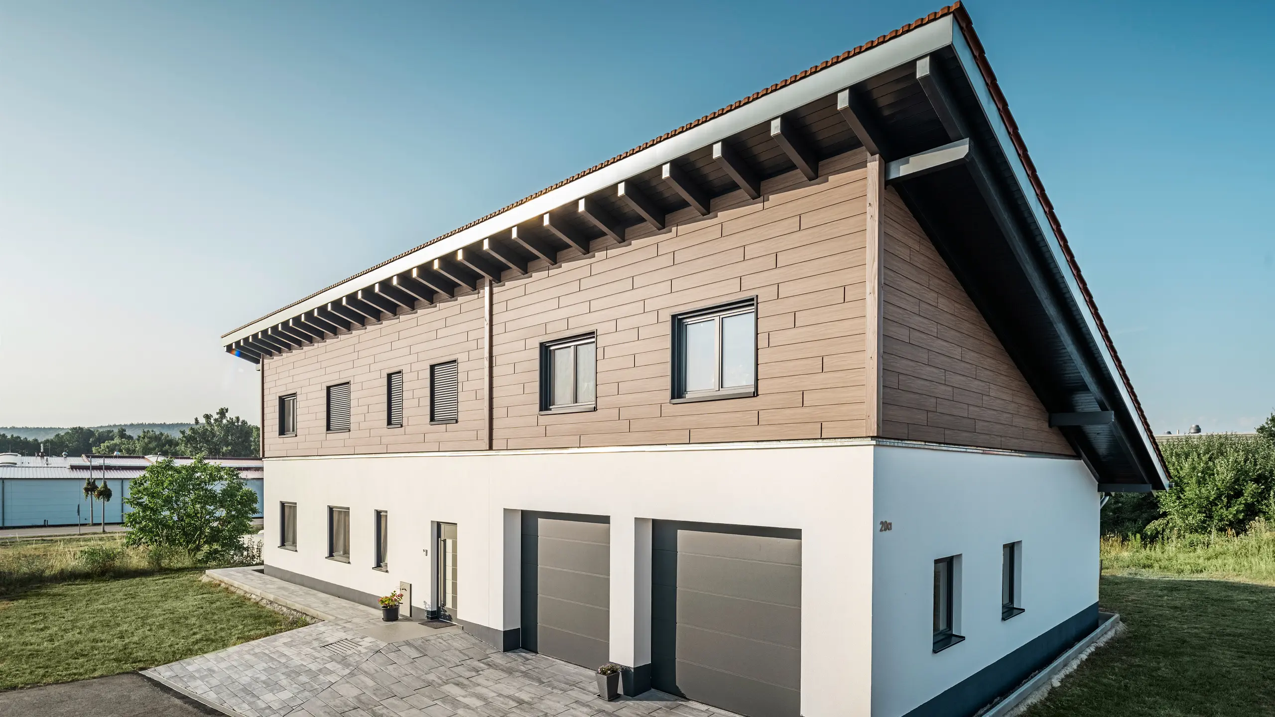 Vy från sidan av en villa med pulpettak i Bruck. Det övre våningsplanet är klätt med PREFA fasadpanel i valnöt. Till höger i bild syns två garageportar i antracit. Entrédörren är placerad i mitten av byggnaden. Sockeln är i en slät vit kulör. 