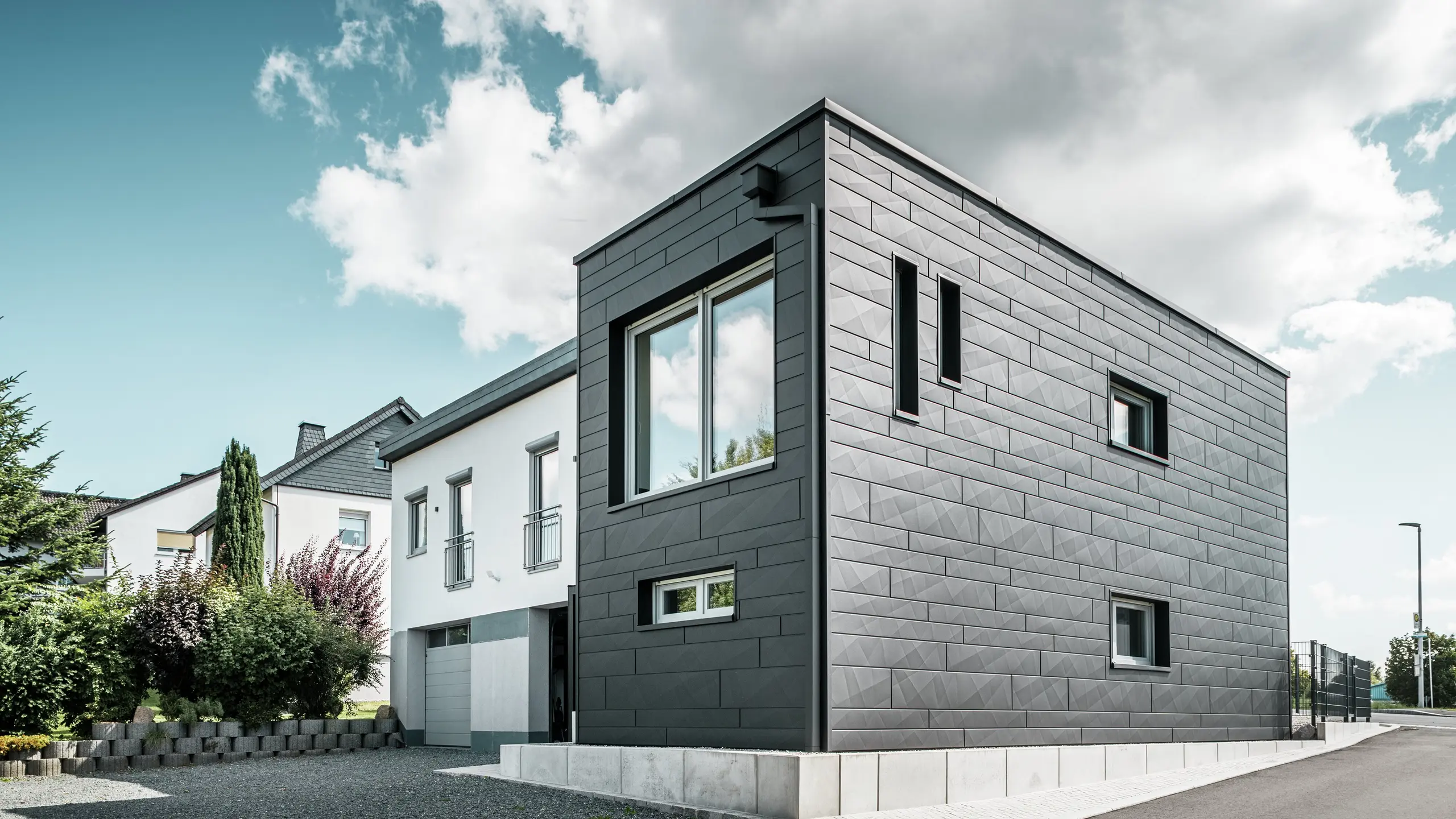 Den stilfulla och moderna fasaden på det renoverade enfamiljshuset i Teuschnitz pryds av de tydligt strukturerade PREFA Fasadpanel.X-panelerna i P.10 svart. Den geometriska placeringen av de högkvalitativa plåtpanelerna ger byggnaden en markant, tidlös estetik. Fönstren är inramade av svarta aluminiumkompositplattor, vilket harmoniskt kompletterar designen. Det integrerade PREFA-takavvattningen med fyrkantrör är en funktionell och estetisk lösning.