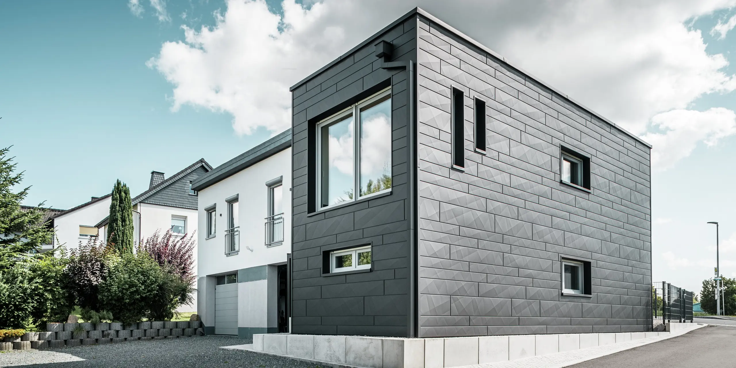 Den stilfulla och moderna fasaden på det renoverade enfamiljshuset i Teuschnitz pryds av de tydligt strukturerade PREFA Fasadpanel.X-panelerna i P.10 svart. Den geometriska placeringen av de högkvalitativa plåtpanelerna ger byggnaden en markant, tidlös estetik. Fönstren är inramade av svarta aluminiumkompositplattor, vilket harmoniskt kompletterar designen. Det integrerade PREFA-takavvattningen med fyrkantrör är en funktionell och estetisk lösning.