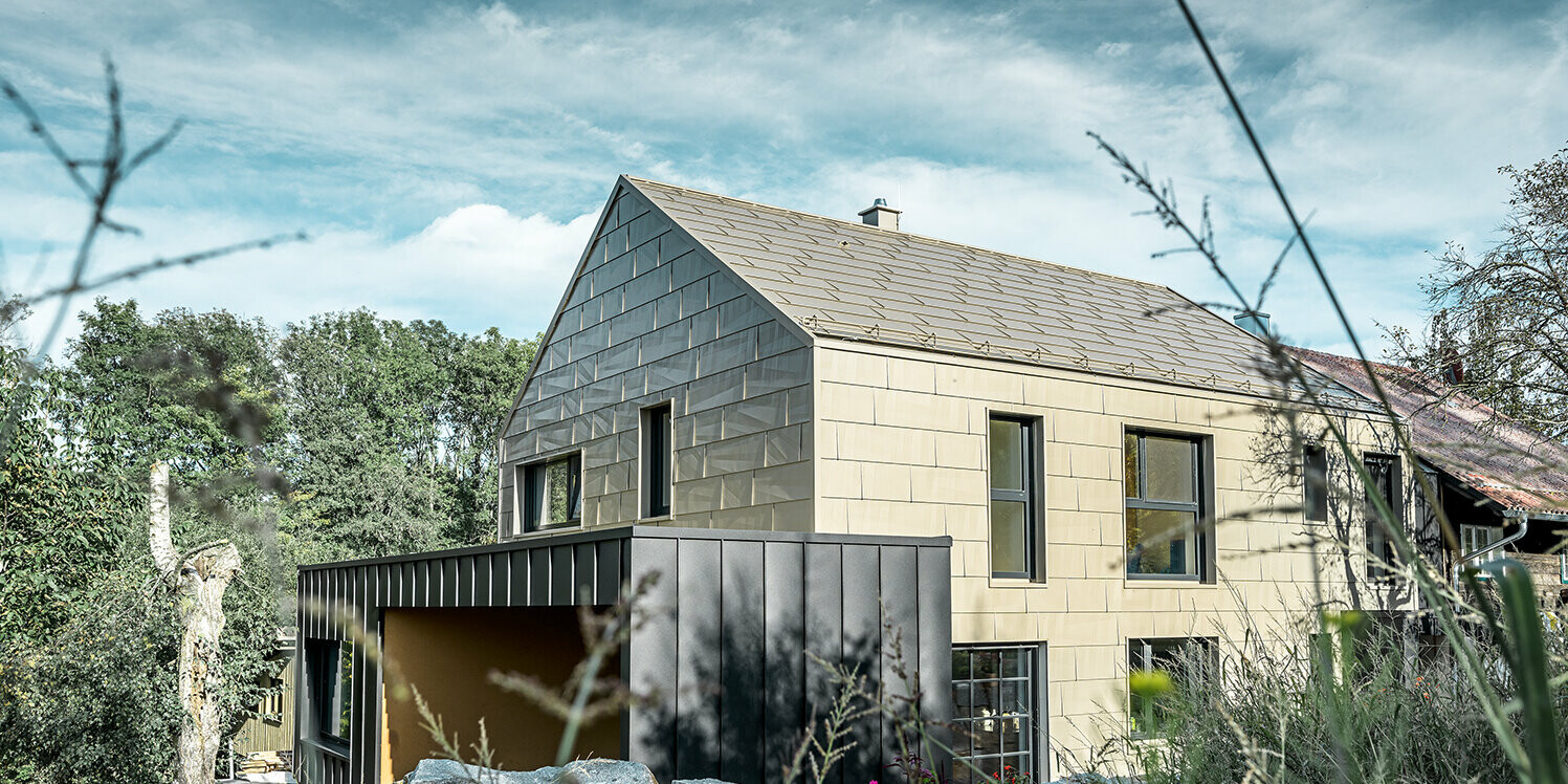Tak och fasad på bostadshus utan taksprång är täckt med aluminiumpanel FX.12 från PREFA i brons. Precis bredvid står ett garage med flackt tak med en fasad med stående vinkelfals.