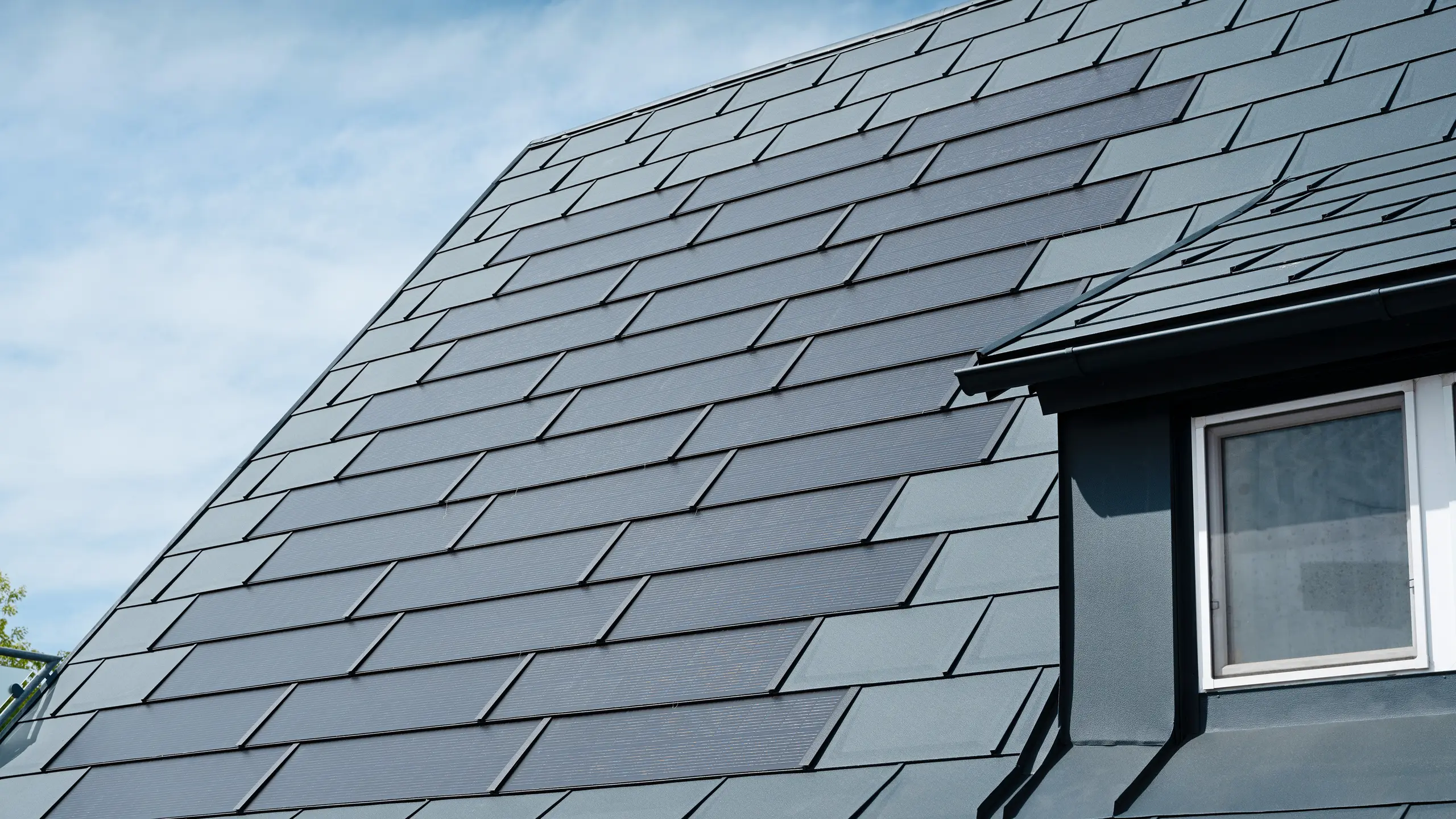 Detaljbild av det renoverade taket på ett enfamiljshus i Leonding med PREFA solar takplatta i antracit. Närbilden visar de högkvalitativa aluminiumpanelerna från PREFA, som möjliggör en sömlös integrering av solmodulerna. Denna hållbara och väderbeständiga lösning erbjuder ett hållbart och energieffektivt sätt att generera elektricitet. Bilden illustrerar den estetiska och funktionella kvaliteten hos de material som används. Till höger i bilden syns ett vitt fönster under en släpande takkupa.